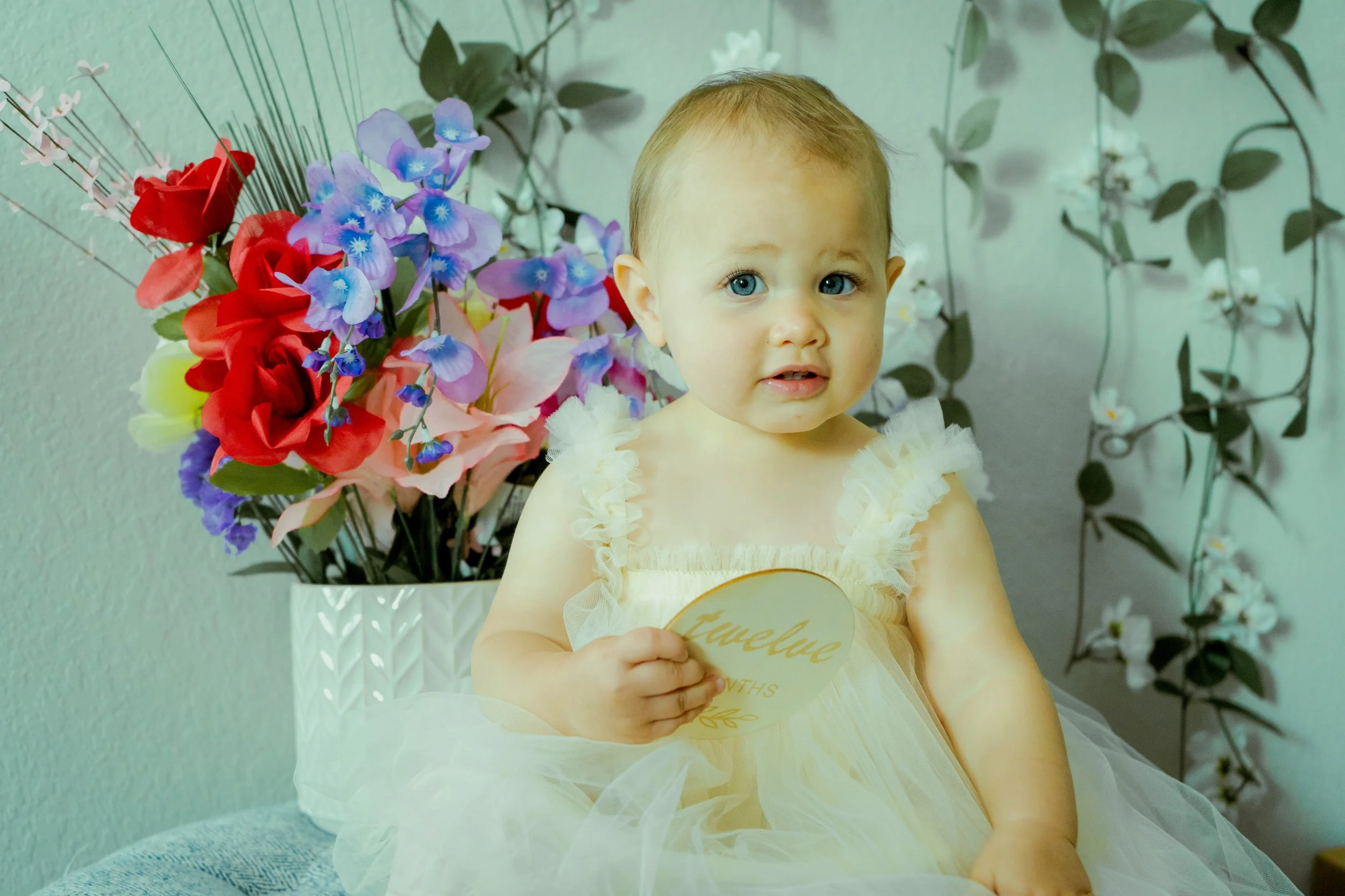 A young girl with blonde hair and blue eyes, wearing a cream-colored dress with ruffled sleeves, holding a round sign with word written on it. She is sitting in front of a large bouquet of colorful artificial flowers, including red, pink, purple, and