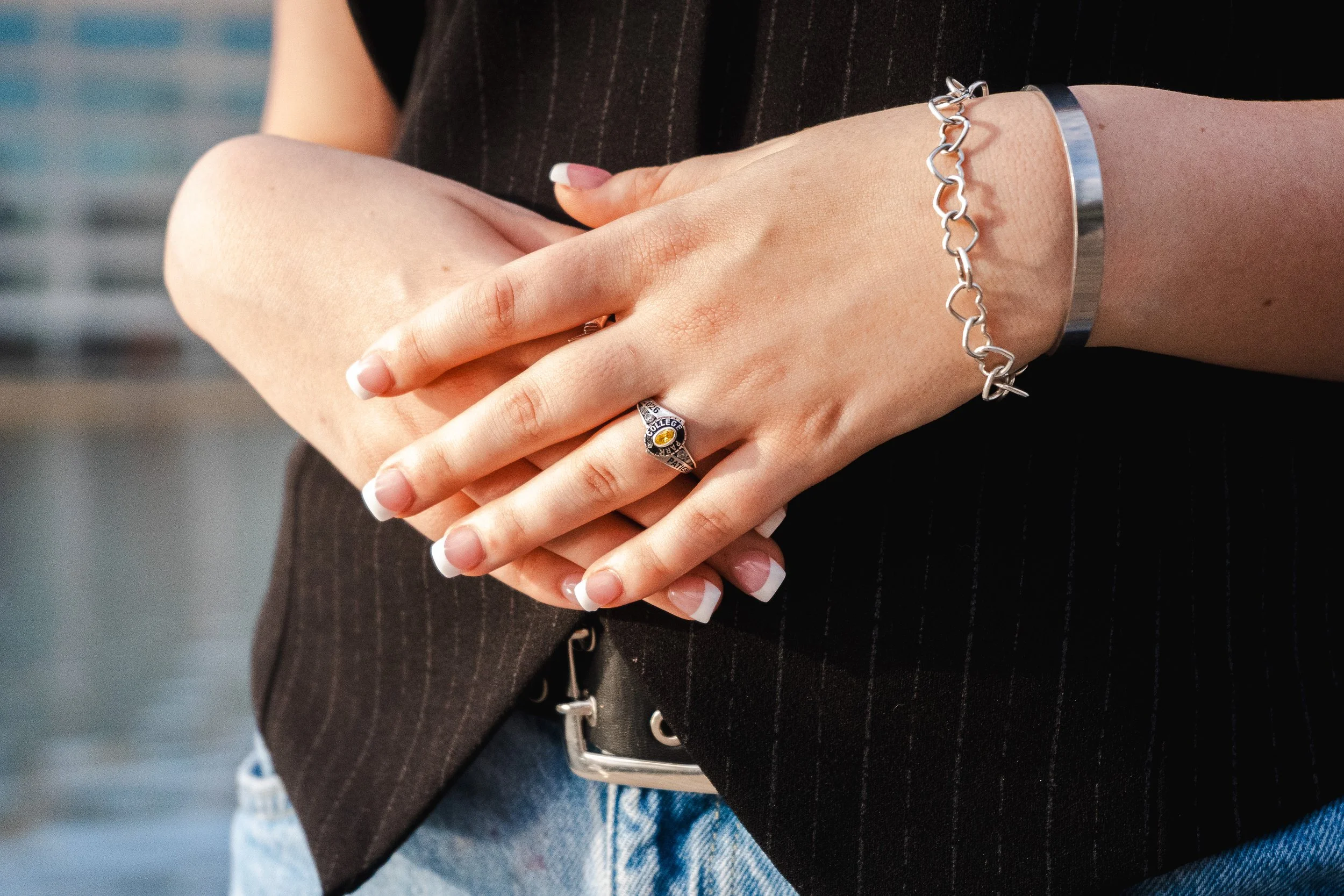 Close-up of two hands intertwined. The person is wearing a silver bracelet, a ring with the words 'College' and 'Graduate', and a dark bracelet. The background is blurred, showing a person wearing a black outfit and blue jeans.
