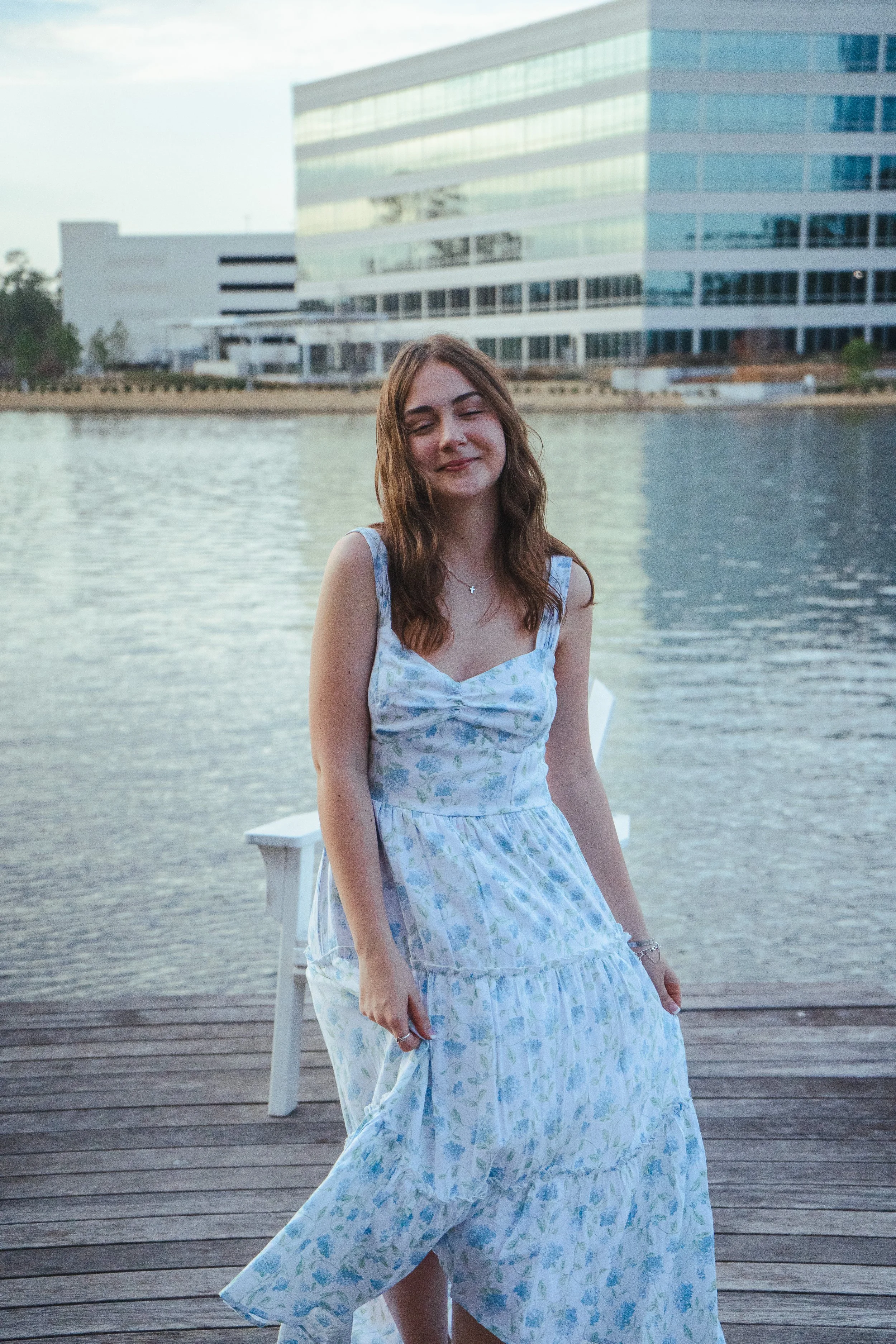 Young woman with long brown hair smiling and wearing a light blue and white floral dress, standing on a wooden dock near water with modern buildings in the background.