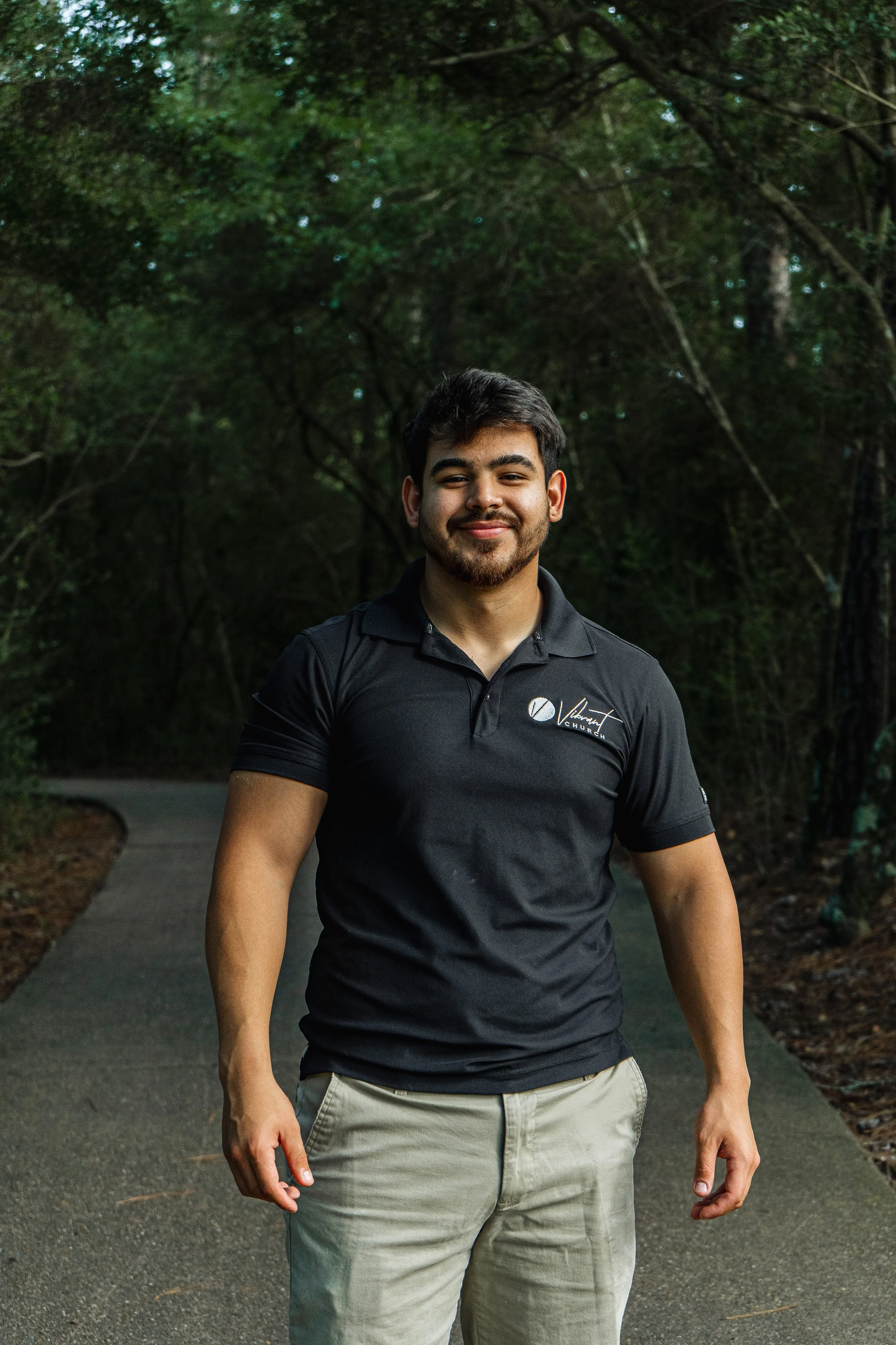 A young man with dark hair, beard, and mustache posing outdoors on a pathway surrounded by trees. He is smiling and wearing a black shirt and beige pants.