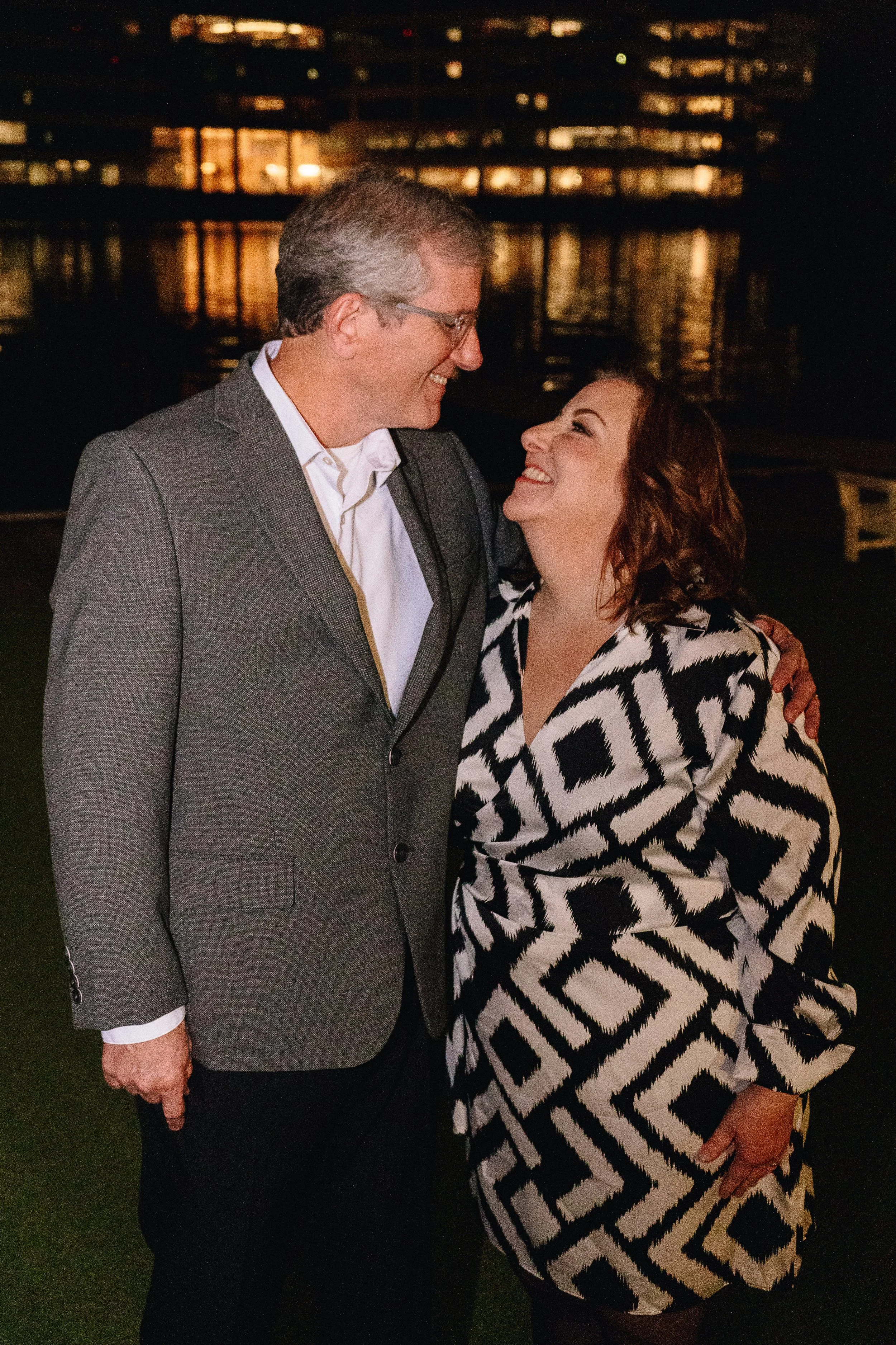 A middle-aged man with gray hair and glasses wearing a gray suit and white shirt, standing close to a woman with dark curly hair wearing a black and white patterned dress, both smiling and looking at each other outdoors at night near a body of water 