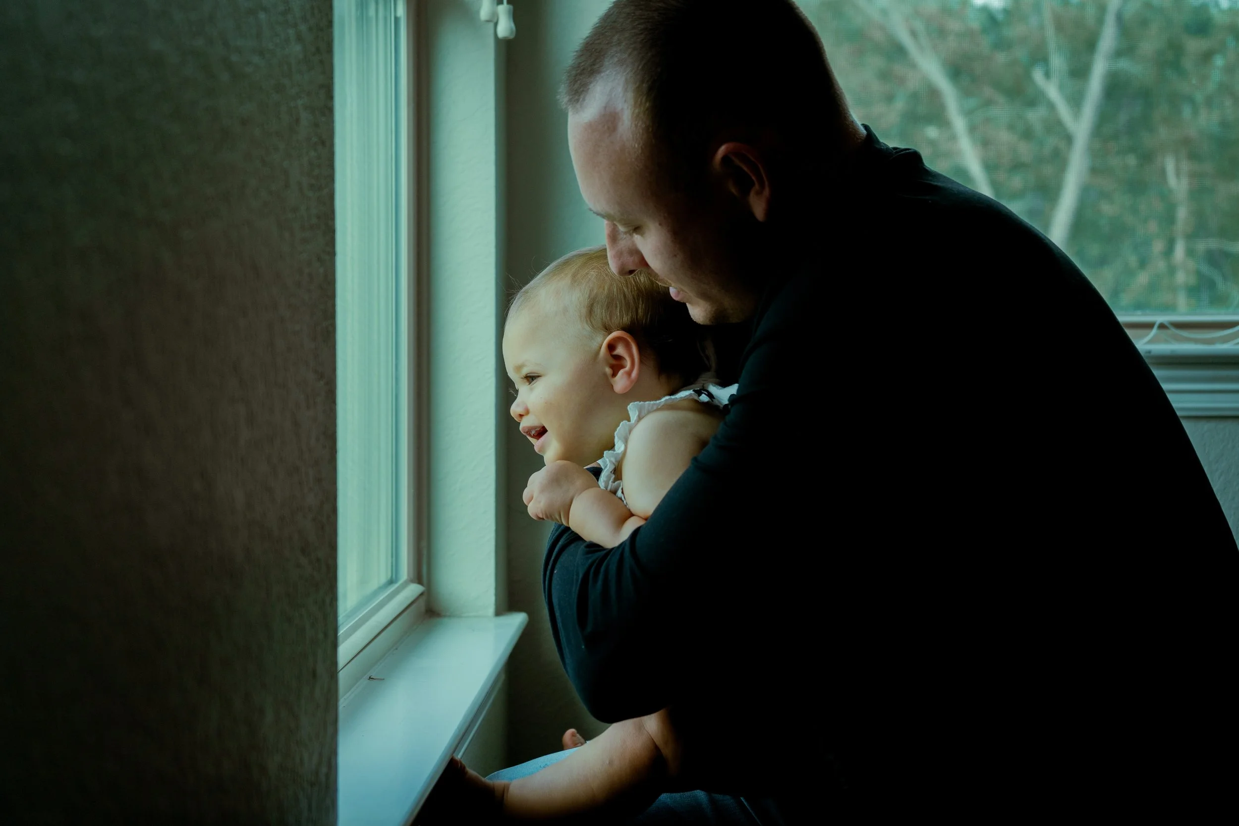An adult man holding a young girl as they look out a window together, with greenery visible outside.