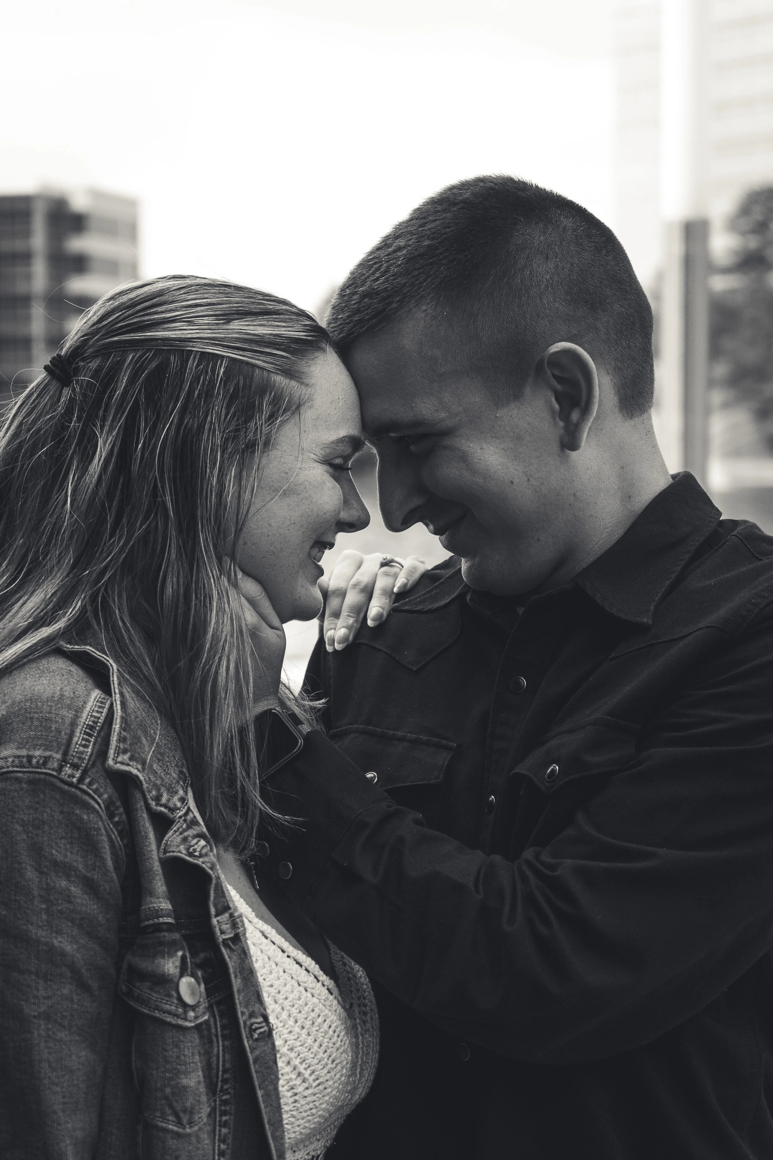 A black and white photo of a couple touching foreheads with their eyes closed, smiling and joyful, outdoors in front of a cityscape.