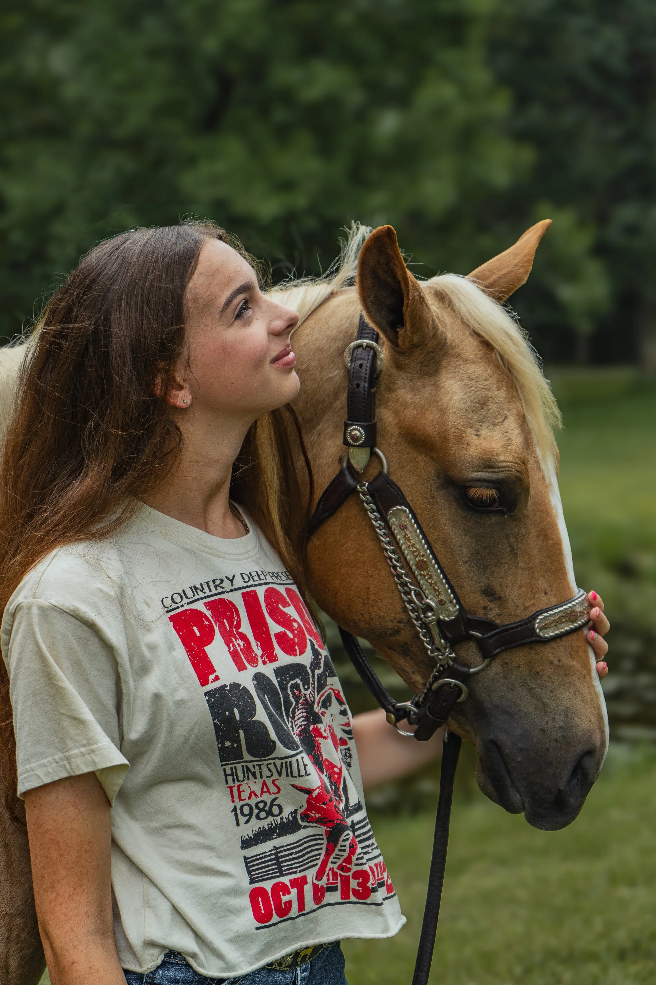 A young woman with long brown hair is standing outdoors, gently holding the head of a light brown horse with a blonde mane. She is wearing a beige T-shirt with red and black text and is looking at the horse's face with affection. The background is bl
