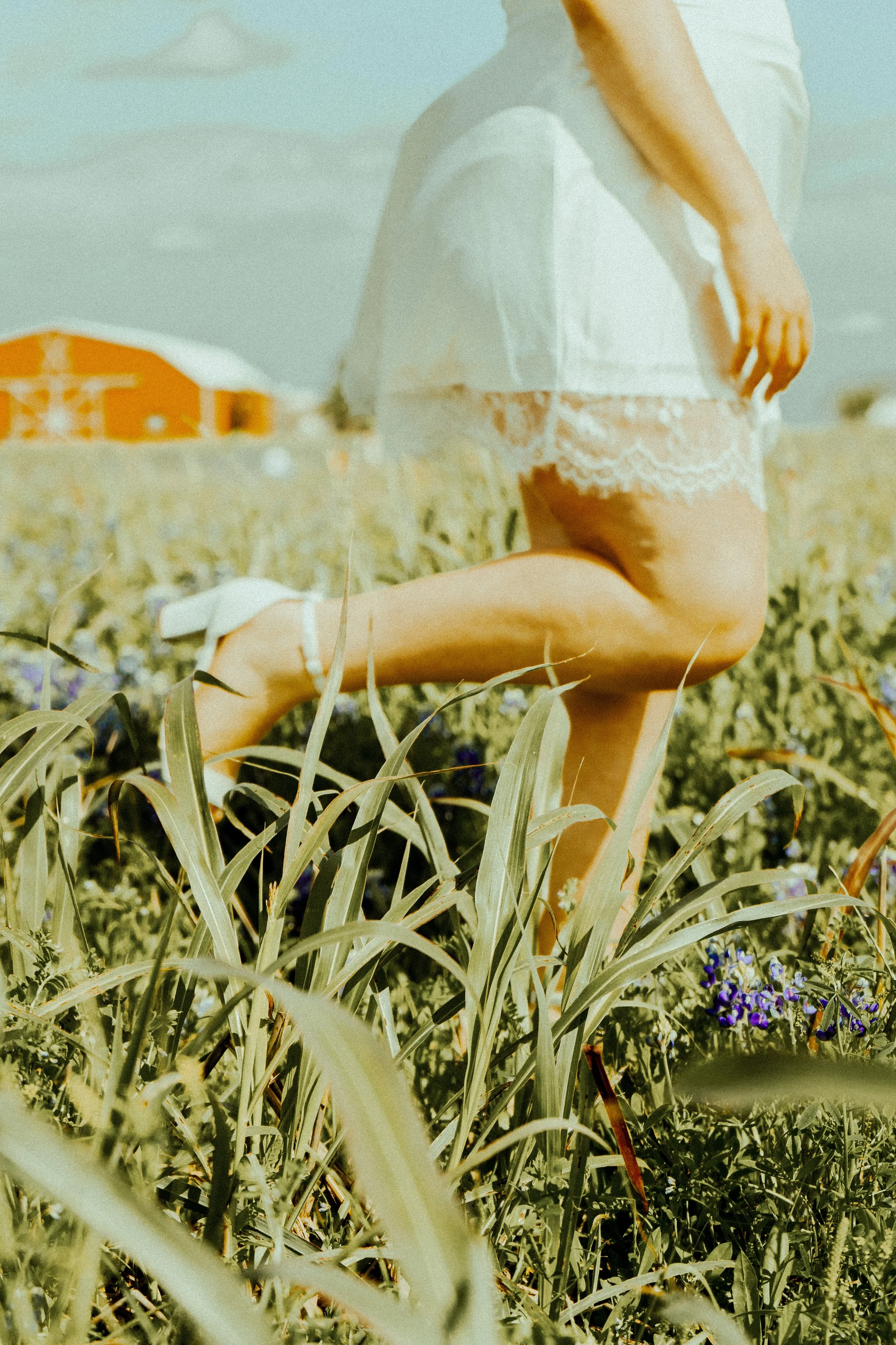 A young woman wearing a white lace dress and a loose white top, kneeling in a field of tall grasses and purple flowers, with a building in the background during daytime.