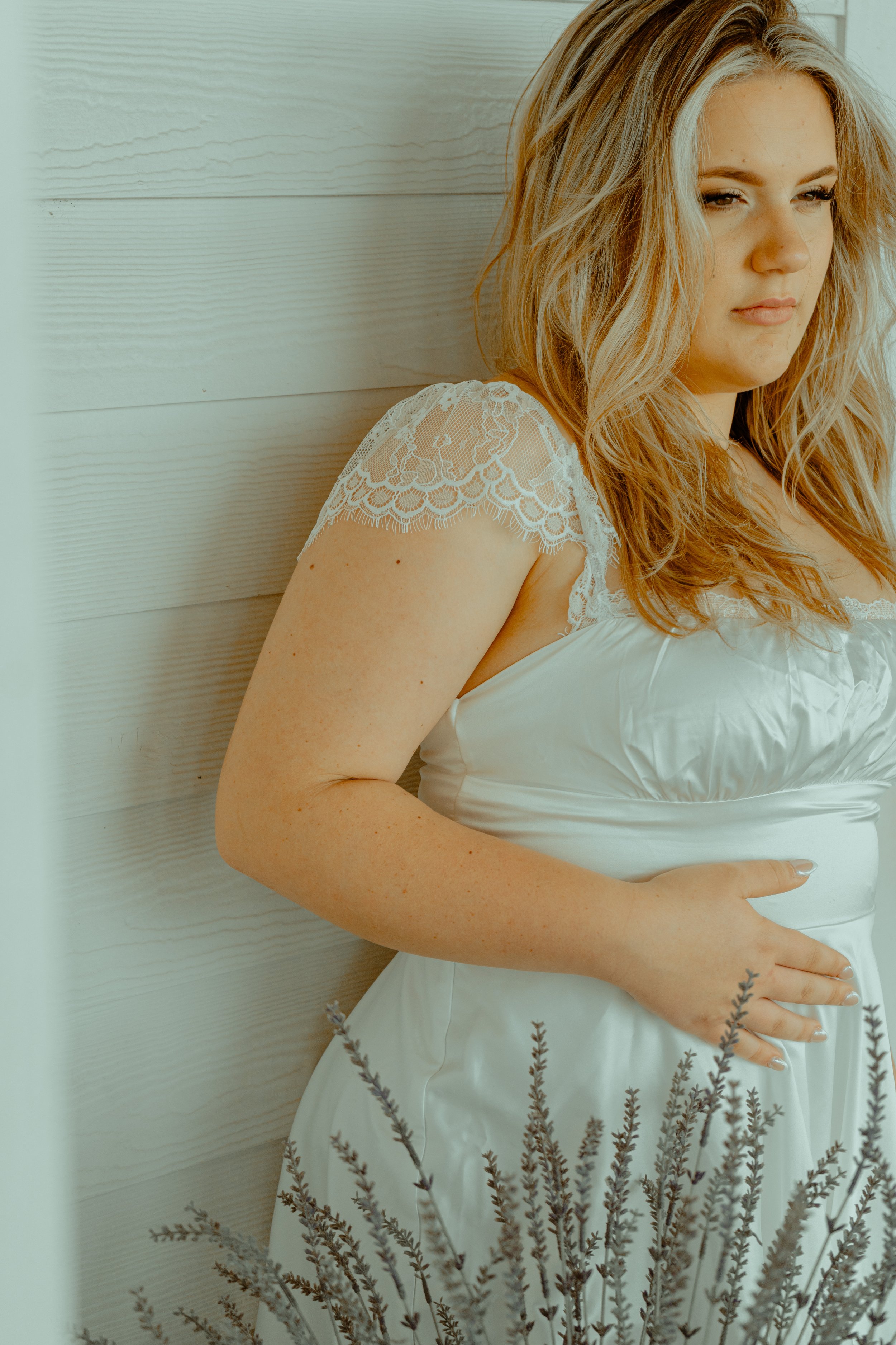 A pregnant woman with blonde hair wearing a white dress with lace sleeves, standing against a white wooden wall, with lavender flowers in the foreground.
