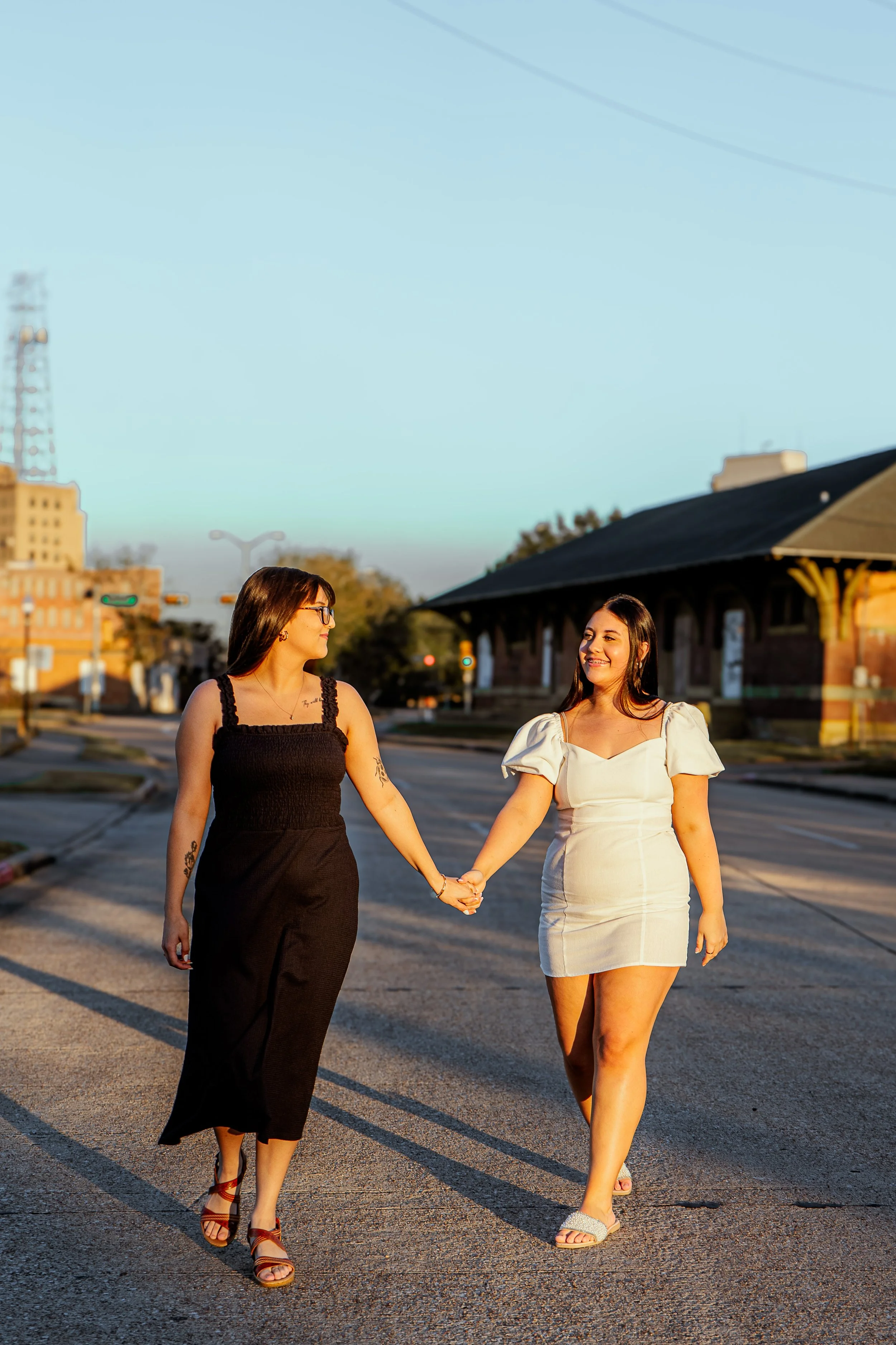 Two sisters holding hands and walking on a city street during sunset, smiling at each other, with buildings and a clear sky in the background.