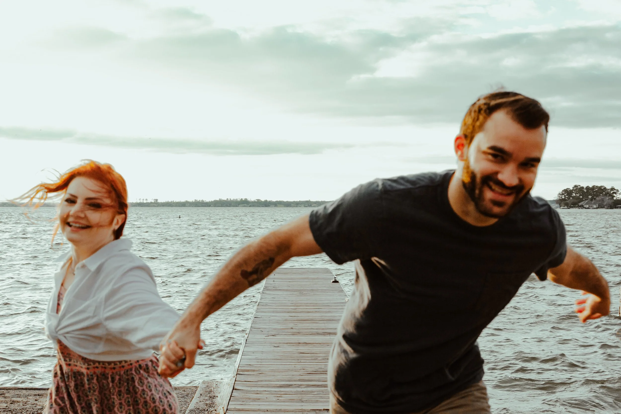 Two people, a woman with red hair and a man with dark hair, are running and holding hands on a wooden dock by the water, with cloudy skies and trees in the background.