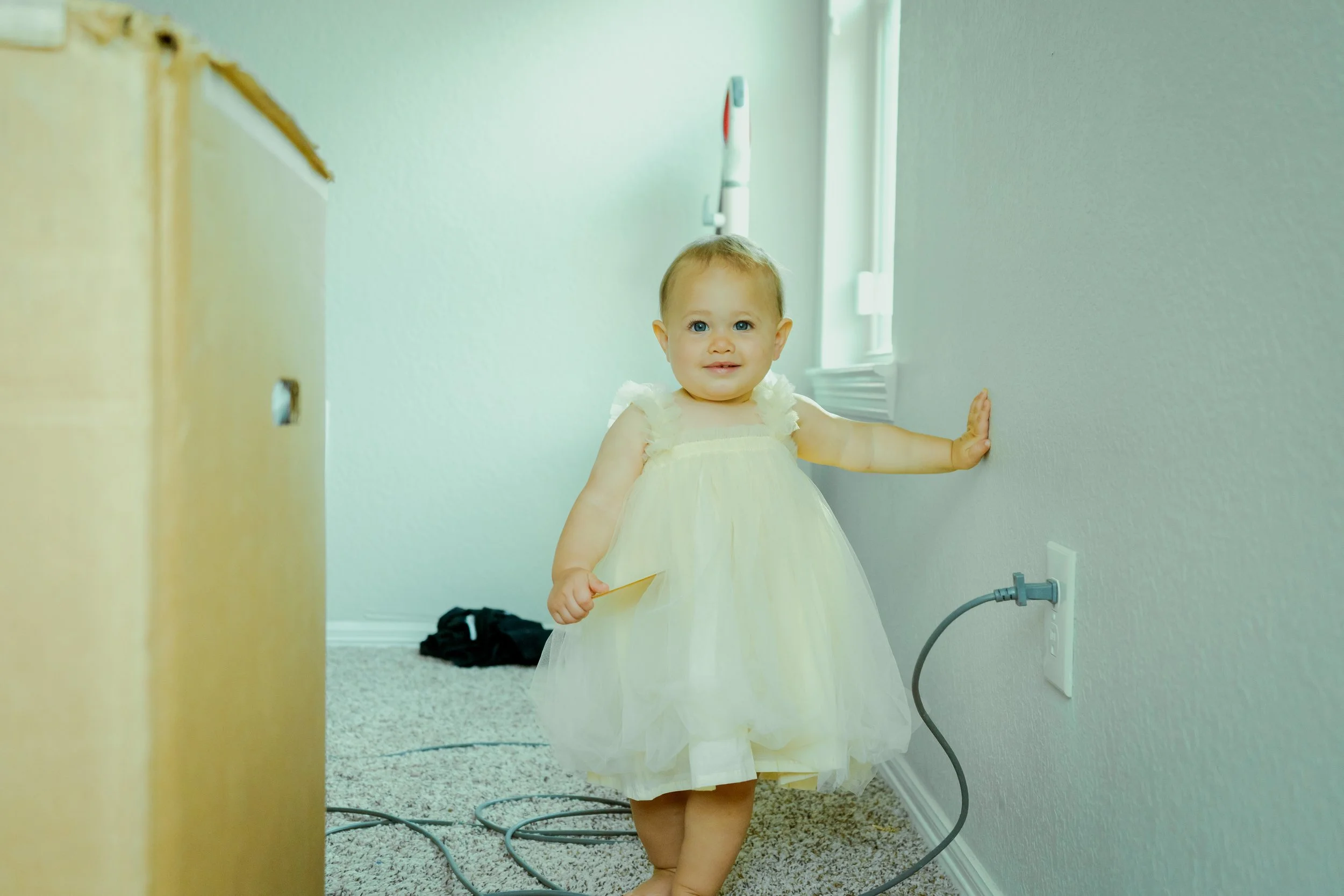 A young girl in a white dress standing in a hallway, touching the wall with her right hand and looking at the camera.