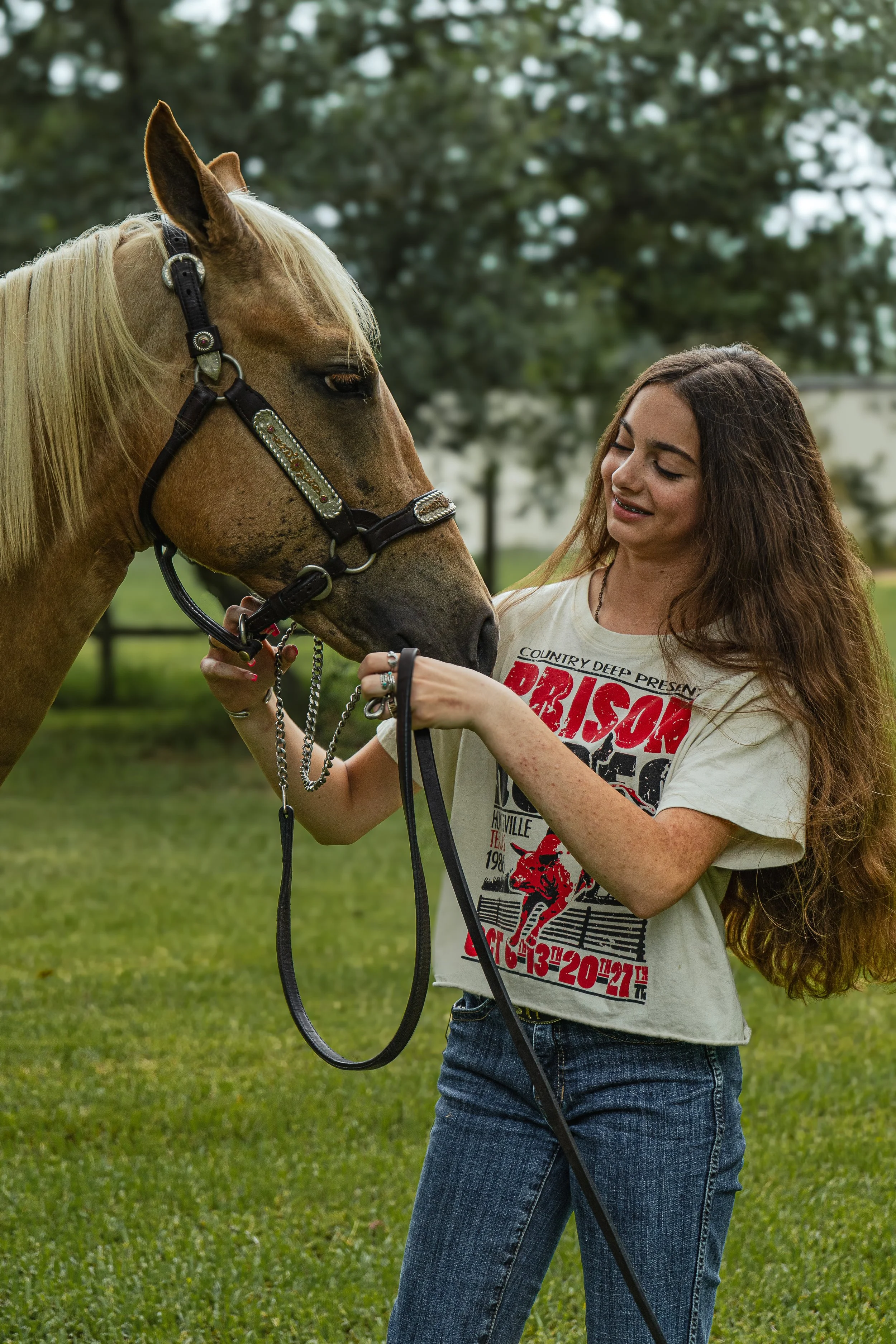 A young woman with long, wavy brown hair and casual clothing smiles as she holds a brown horse by its bridle in a grassy outdoor setting with trees in the background.