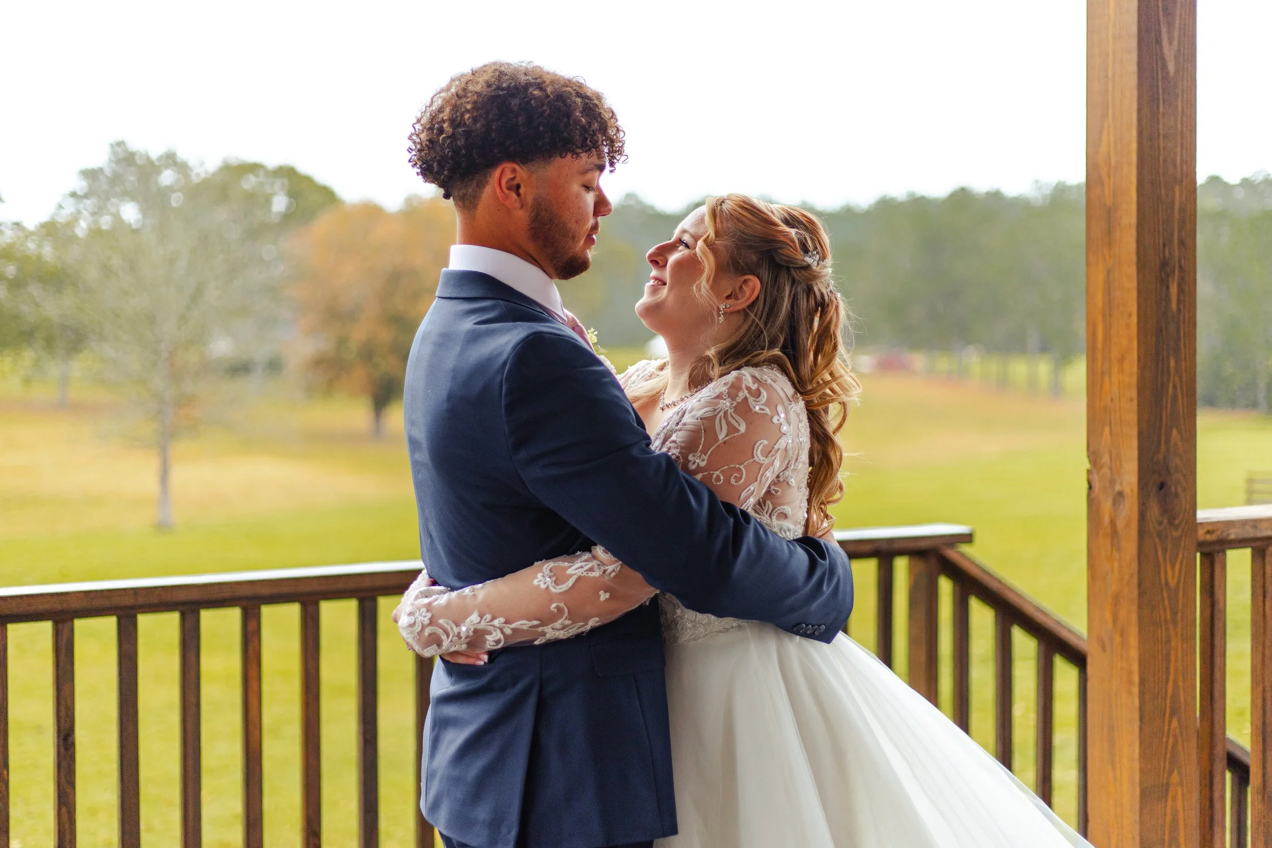 A couple dressed in wedding attire embracing on a wooden porch with trees in the background.