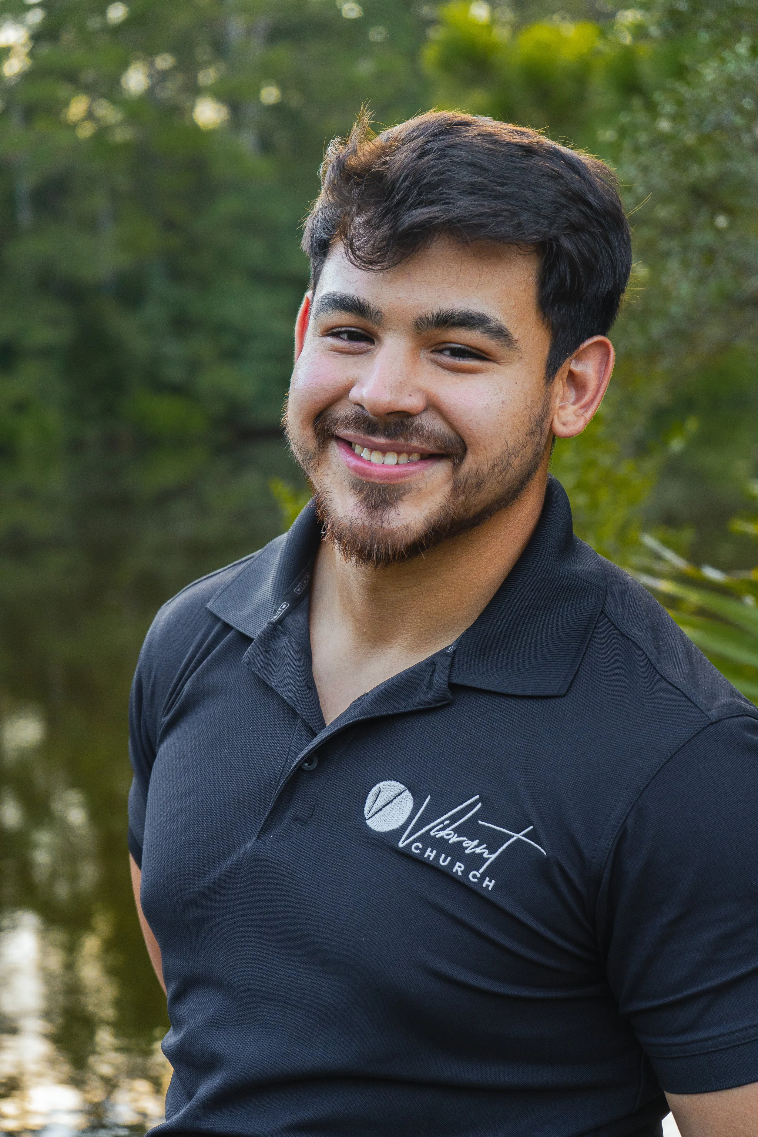 Portrait of a young man with dark hair and a beard, smiling outdoors in front of a blurred background of green trees and water. He is wearing a black polo shirt with a logo that reads "Vibrant Church."