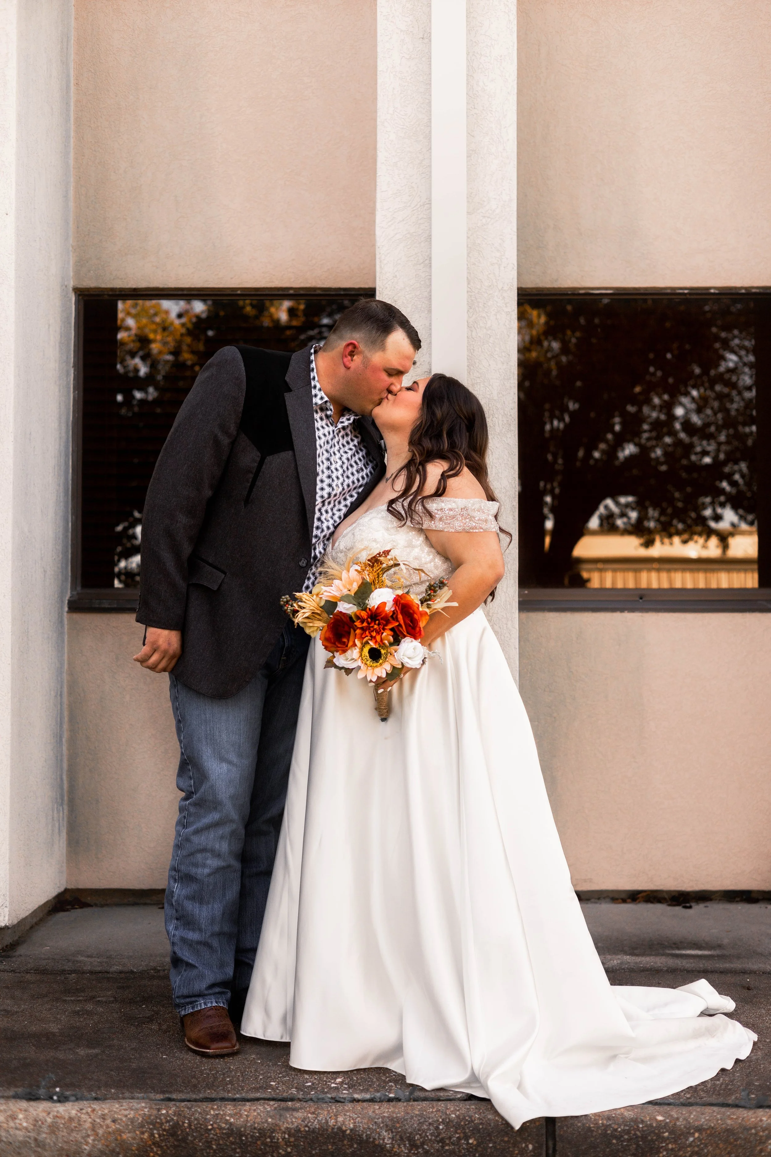 A couple dressed in wedding attire sharing a kiss outdoors next to a building, holding a bouquet of orange, yellow, and white flowers.