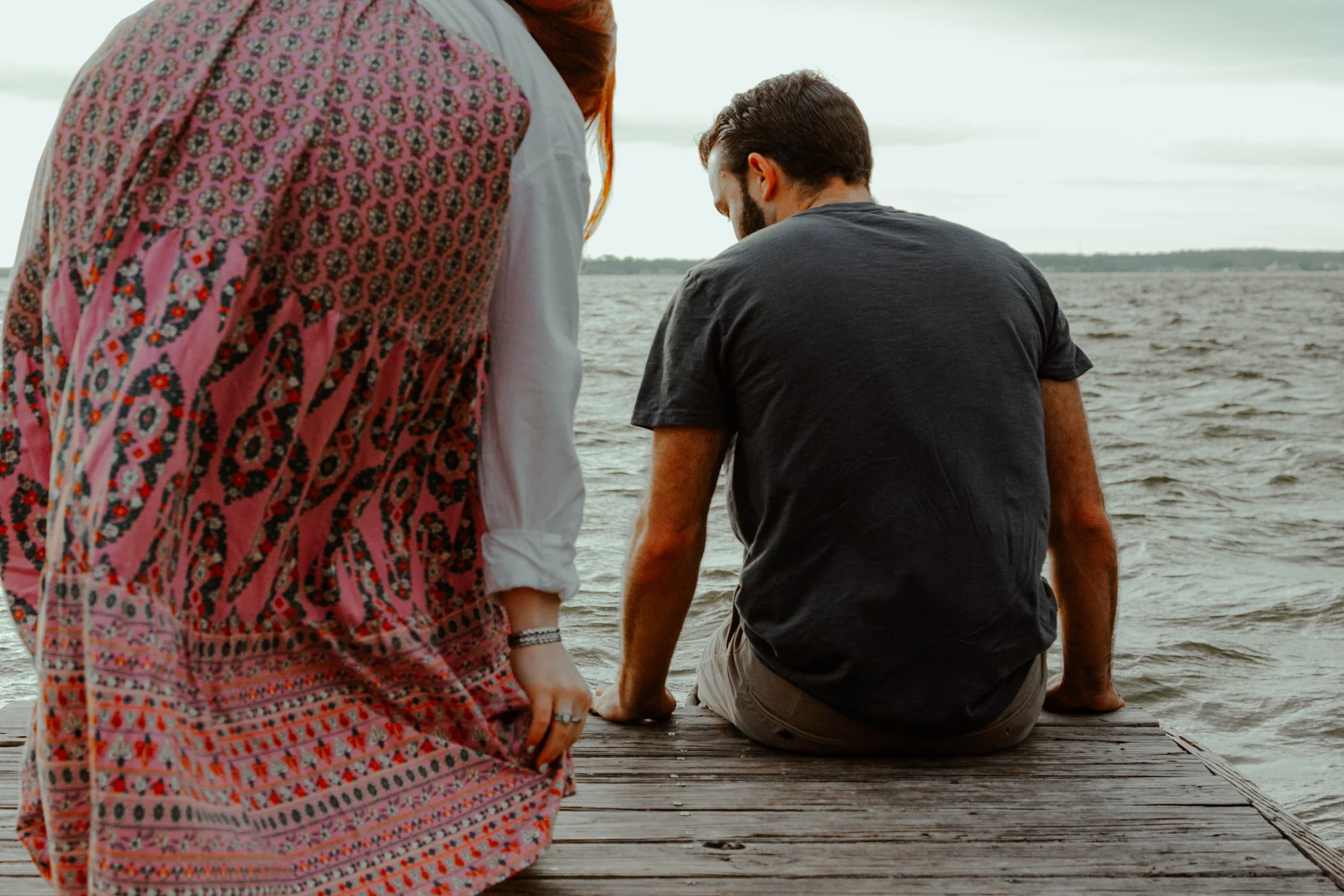 A woman in a pink patterned dress standing on a wooden dock, looking at two men sitting by the water, with a lake and cloudy sky in the background.