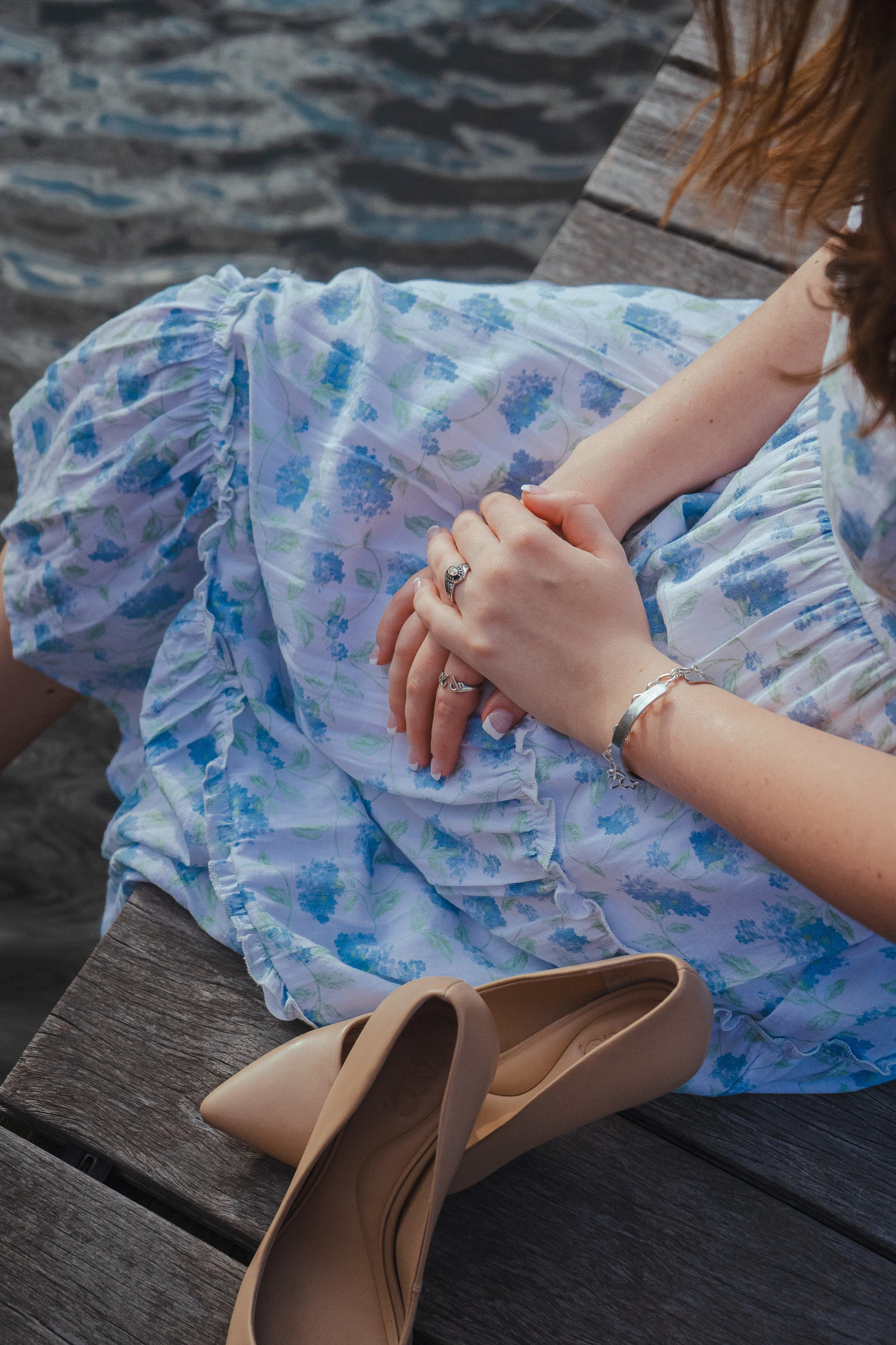 Close-up of a woman sitting on a wooden bench, wearing a floral dress, with hands resting on her lap. Tan high heel shoes are placed on the bench beside her, against a background of a textured wooden surface and a body of water.