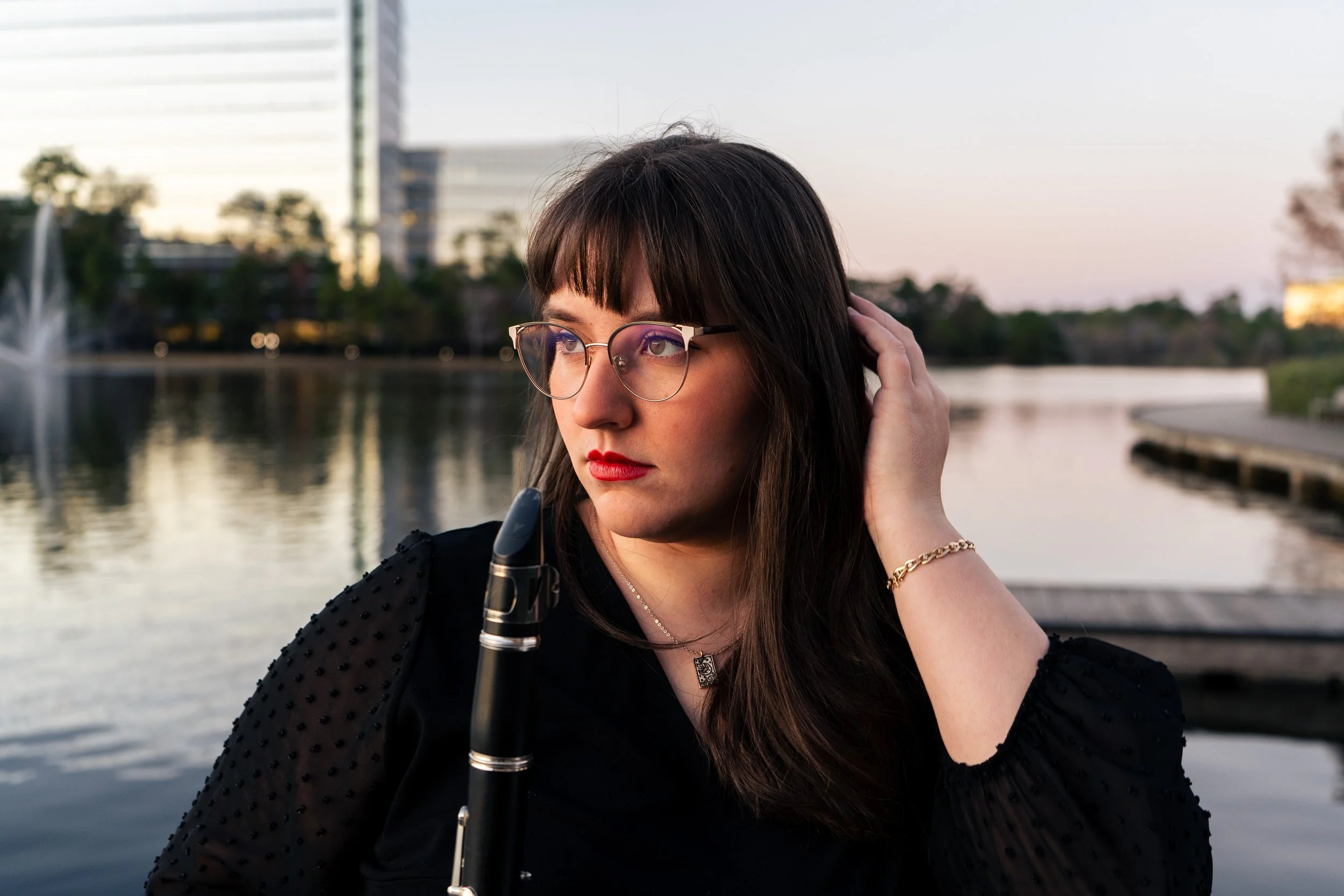 A woman with dark hair and glasses holding a flute, standing near a body of water with modern buildings and trees in the background during the evening.