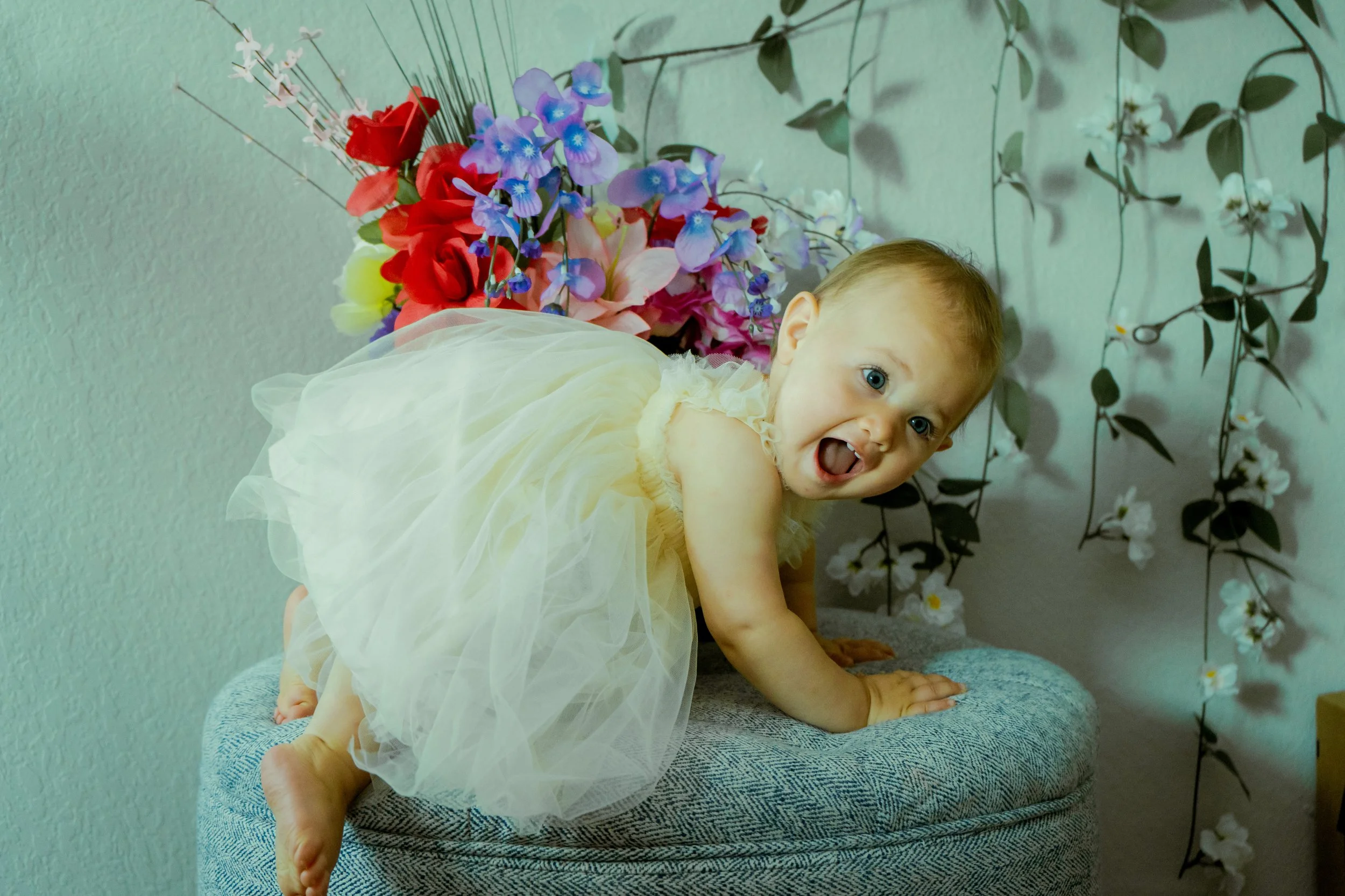 A young child with light skin and blue eyes wearing a light yellow dress with tulle, crawling on a gray cushion with a green wall background decorated with 'Fall' themed flowers and leaves.