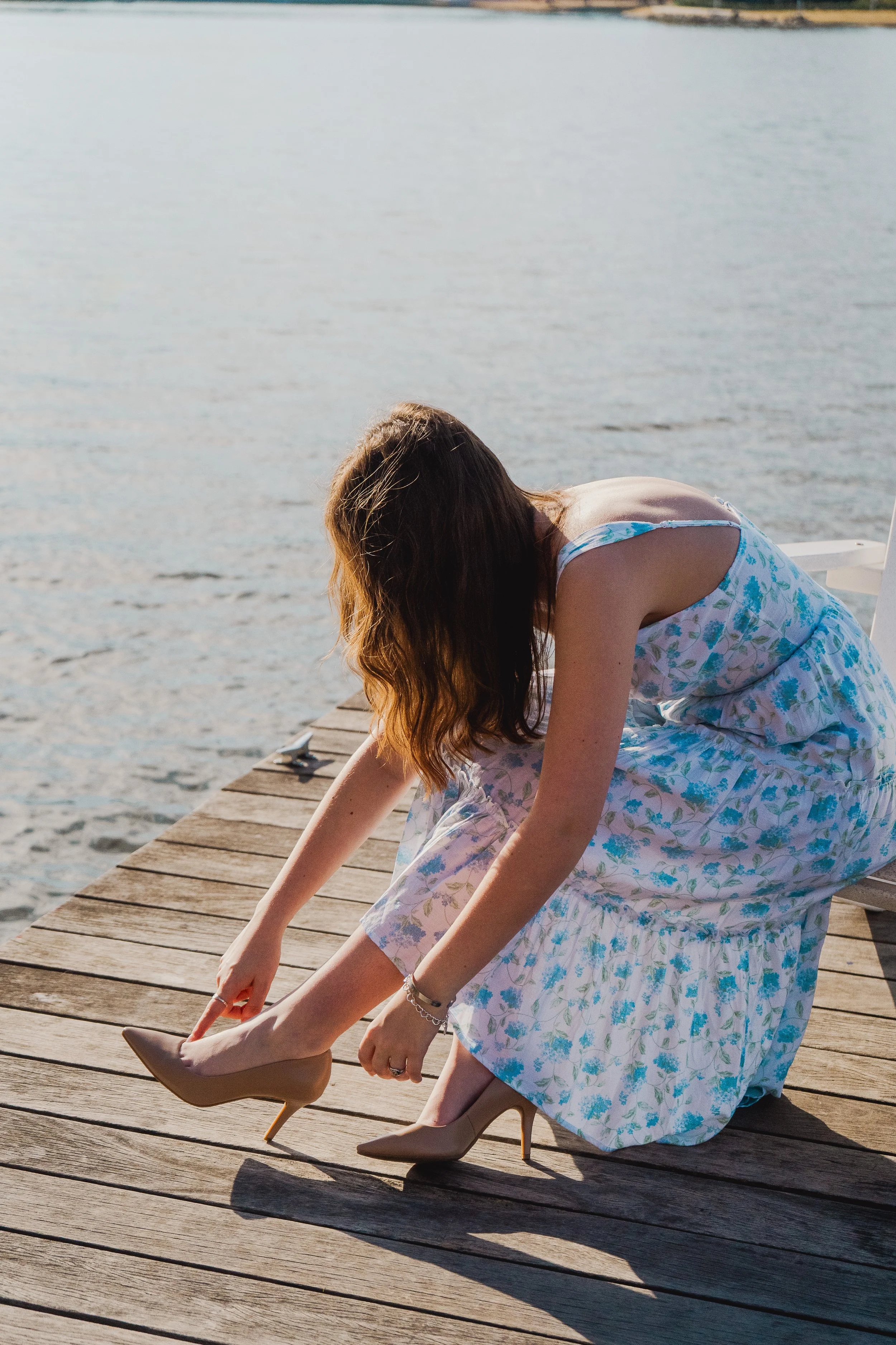 A woman with long, wavy hair wearing a floral dress and nude high heels, sitting on a wooden dock by a body of water during daytime.