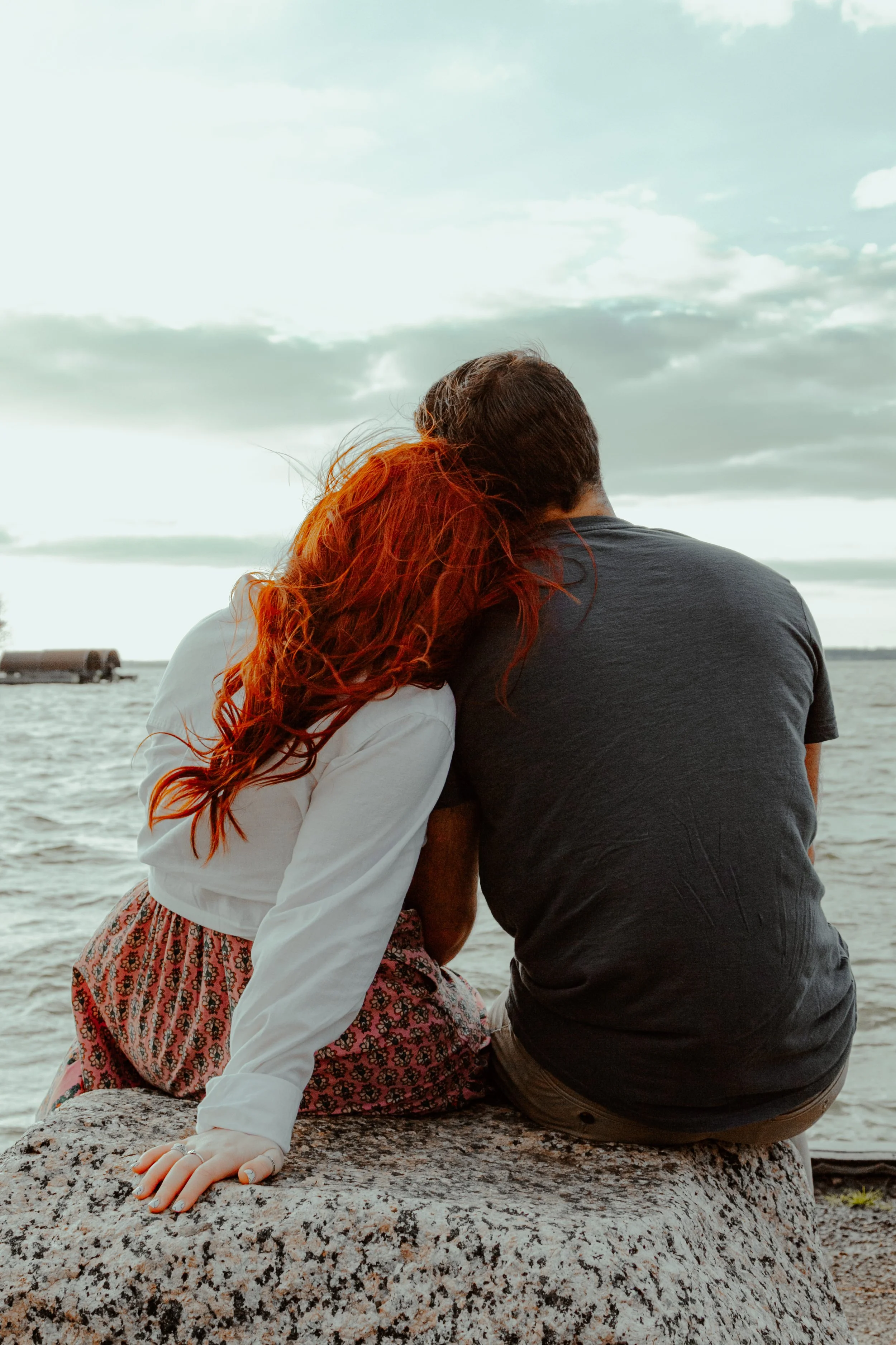 A couple sitting on a rock by the water, embracing each other with their heads touching, facing the sky and clouds.