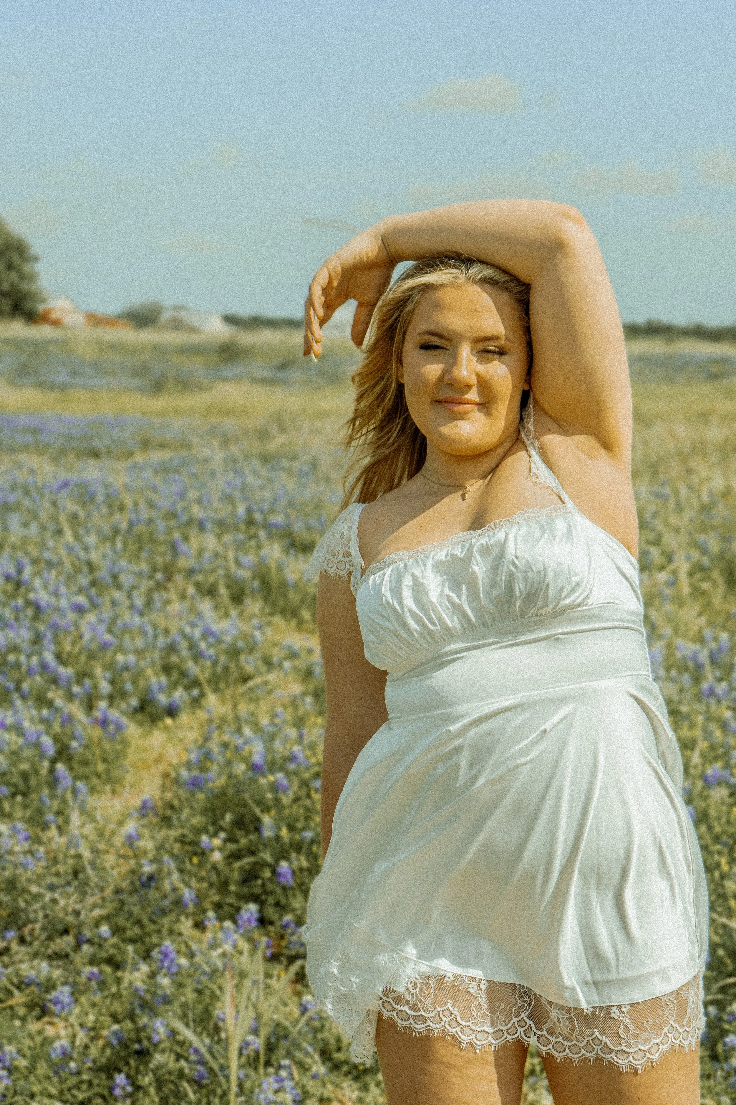 A woman with blonde hair wearing a white dress standing in a field of purple flowers on a sunny day.