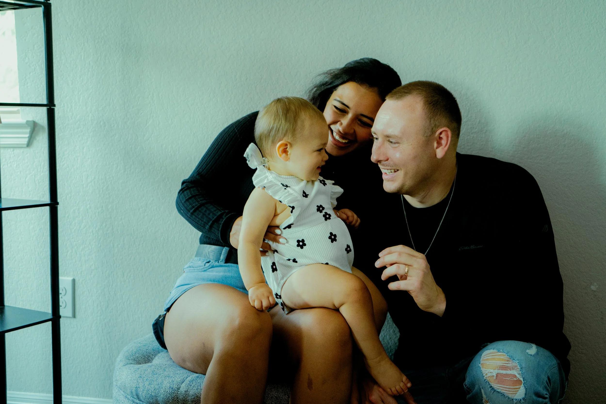 Three people, a woman, a man, and a young girl, are sitting together in a room. They are smiling and looking at each other affectionately. The woman and the man are sitting close to each other, while the girl is sitting on the woman's lap, and they a