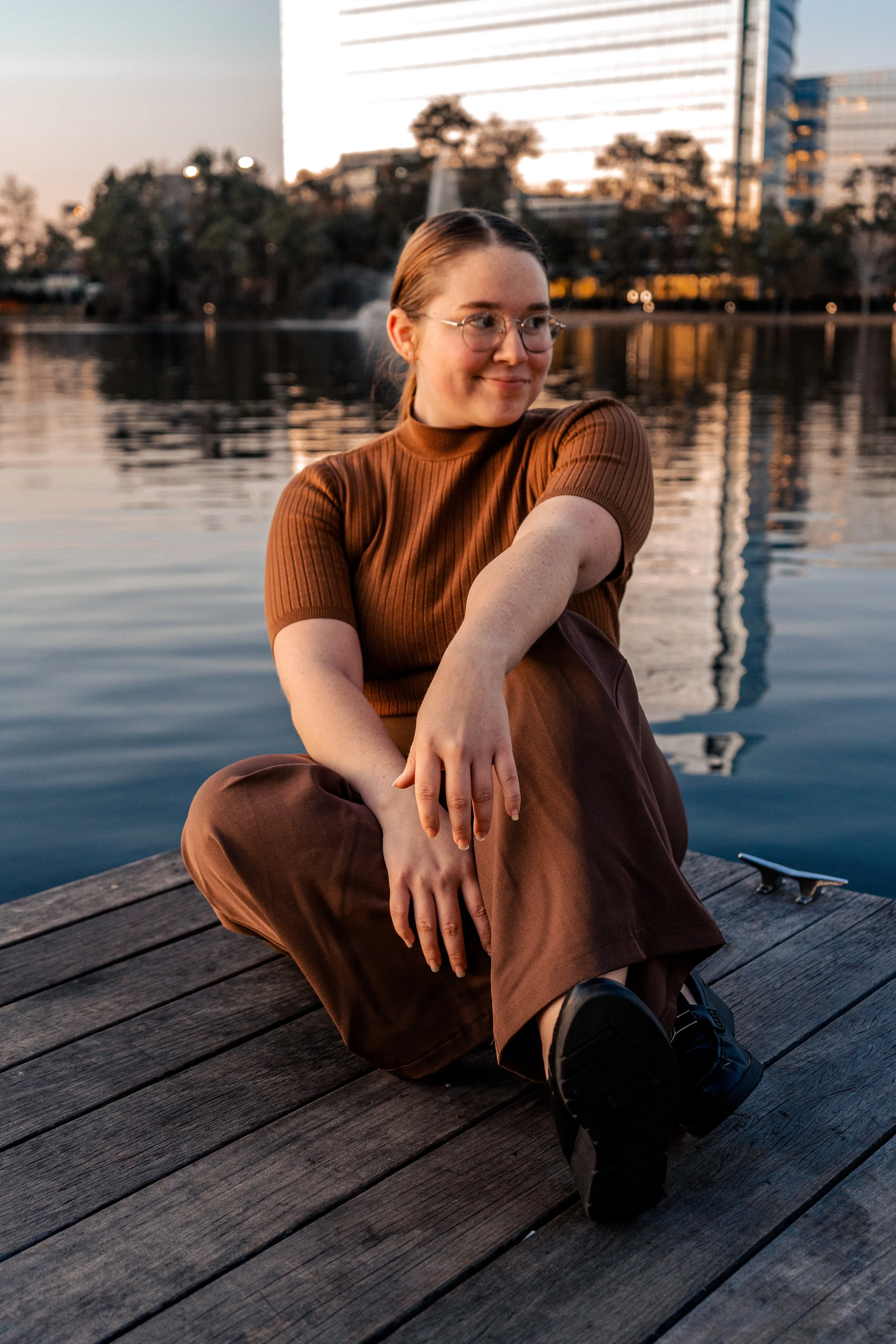 A young woman with glasses and brown hair tied back, sitting on a wooden dock by a lake during sunset, wearing a brown ribbed short sleeve top and brown pants, with a skateboard nearby.
