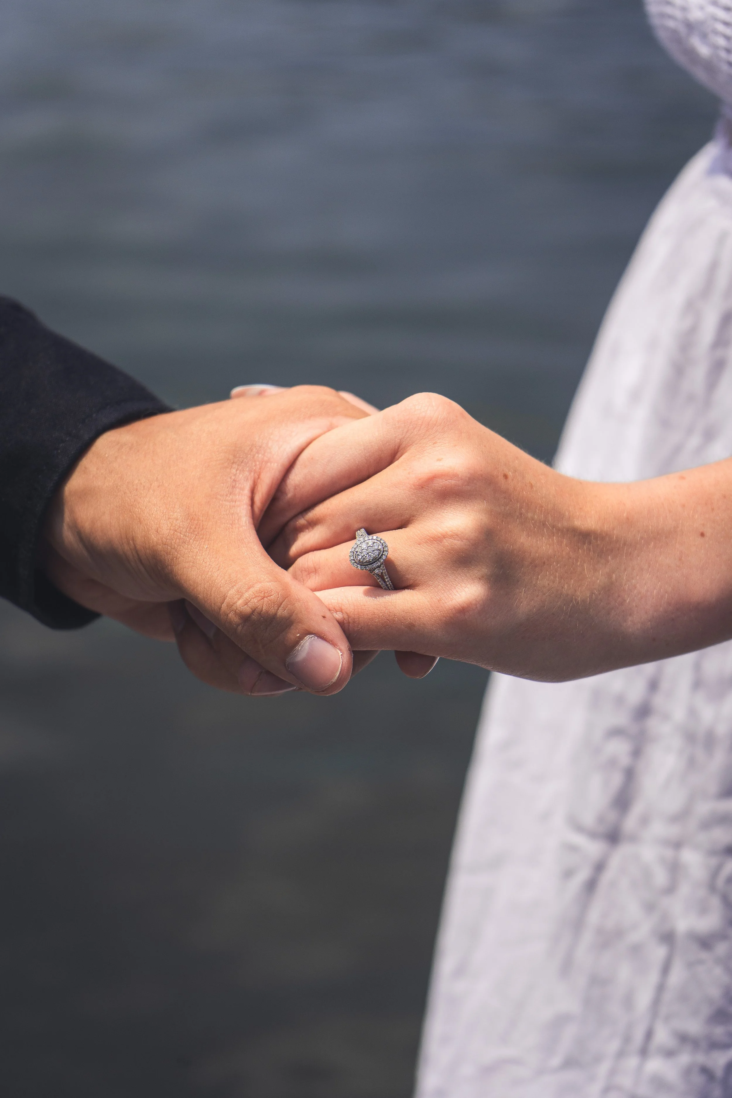 Close-up of a couple holding hands with a wedding ring, against a blurred water background.