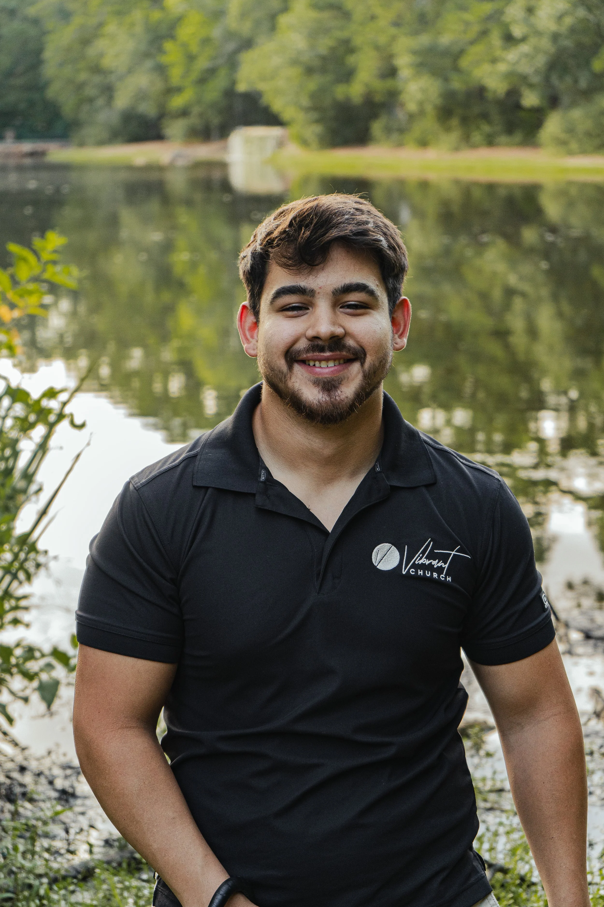 A smiling young man with dark hair and a beard, wearing a black polo shirt with a logo, standing outdoors near a body of water with trees in the background.