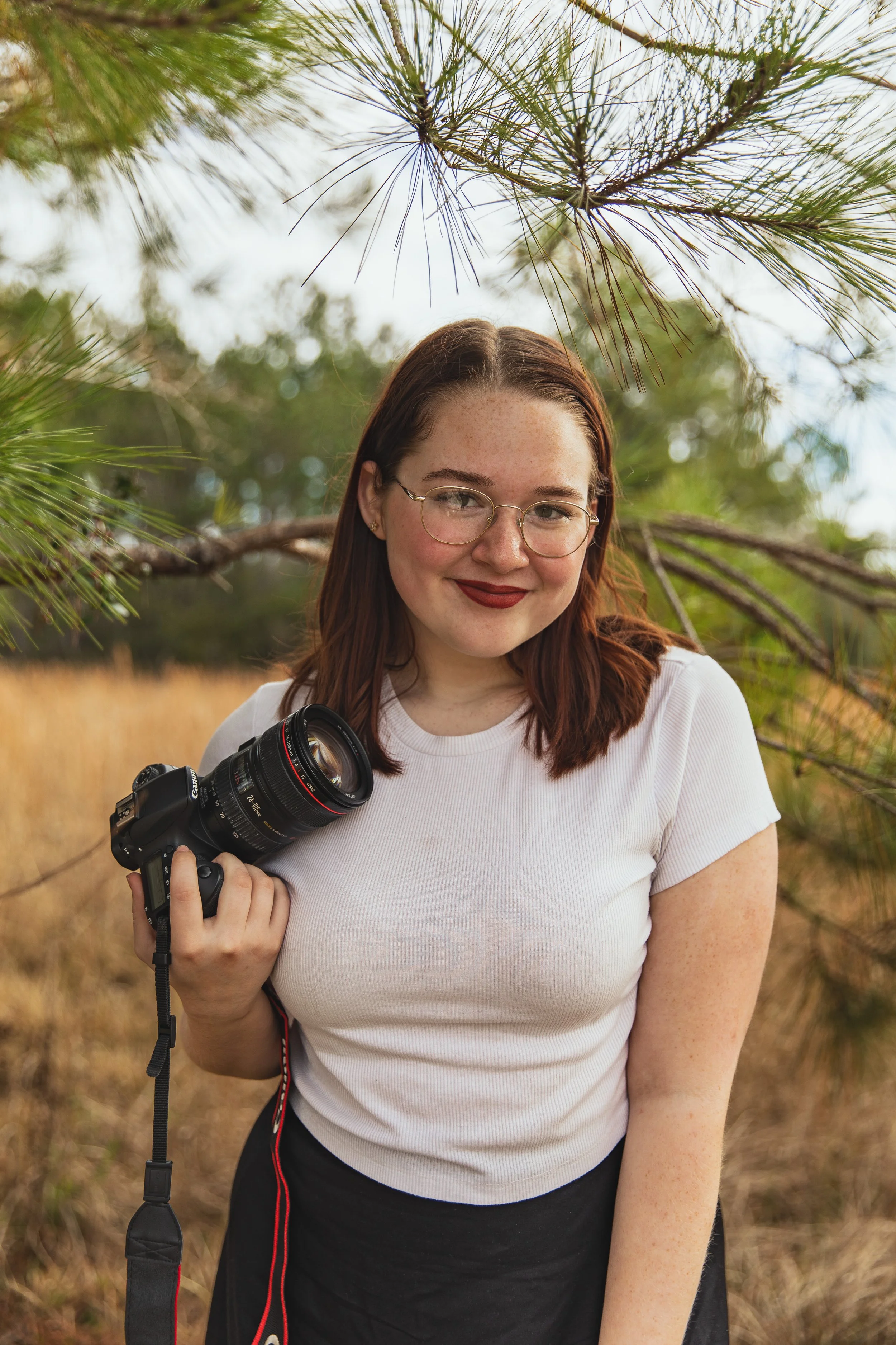 A young woman with red hair and glasses holding a camera outdoors near pine trees and a field.