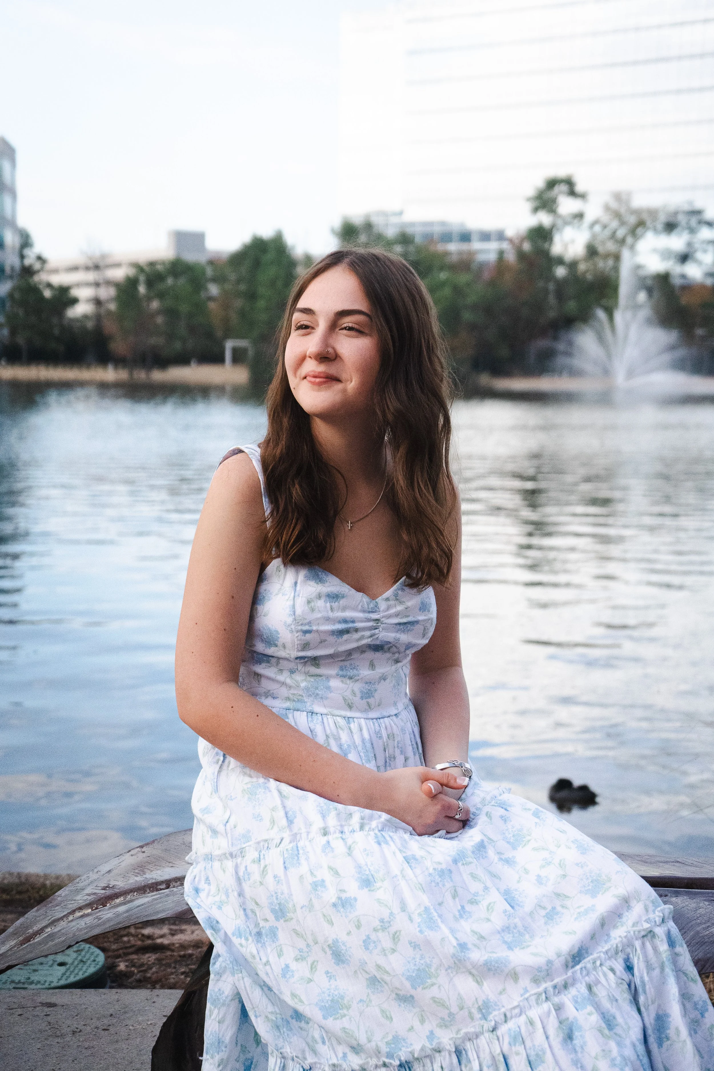 A young woman with long brown hair, wearing a white and pastel floral dress, sitting on a rock by a lake with trees and city buildings in the background, during daytime.