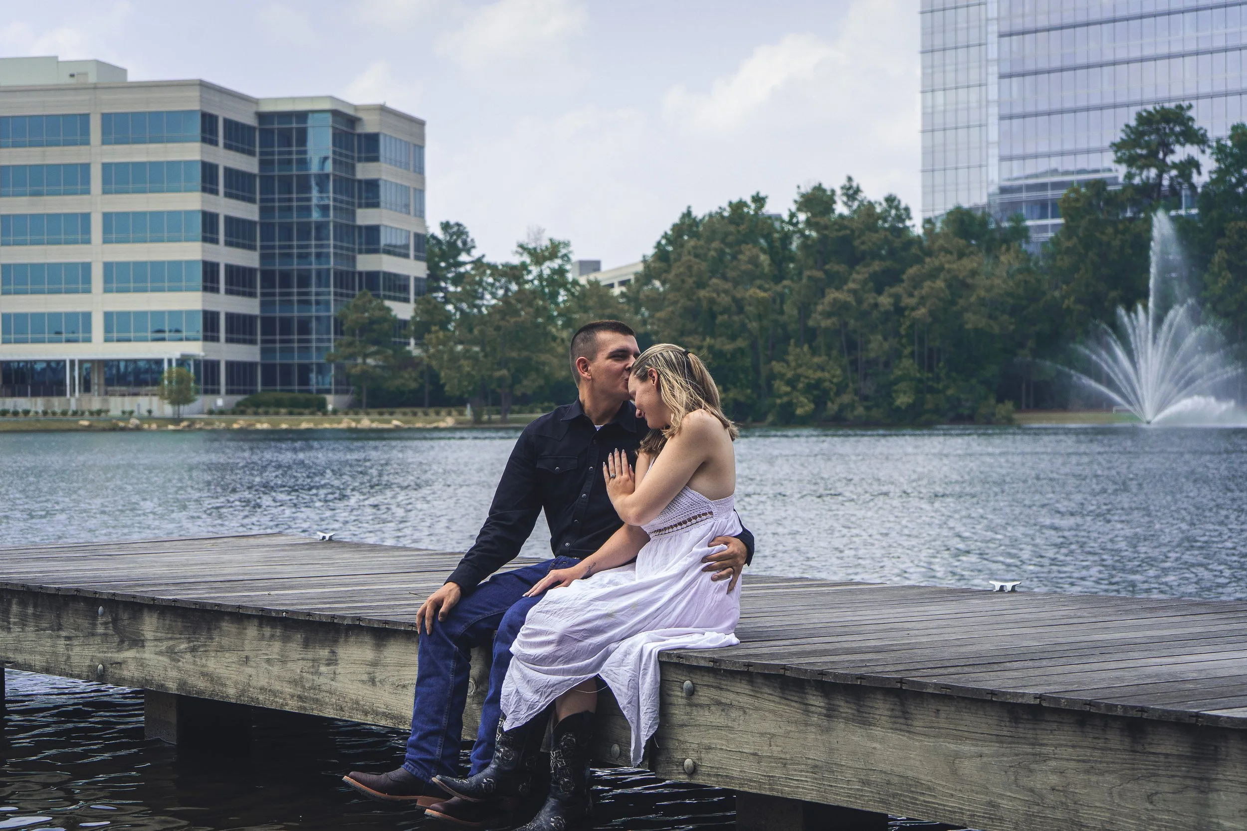 A couple sits on a wooden dock by a lake in an urban park. The man is kissing the woman's forehead while holding her close. The woman is wearing a white dress, and the man is dressed in a dark shirt and jeans. In the background, there are modern glas