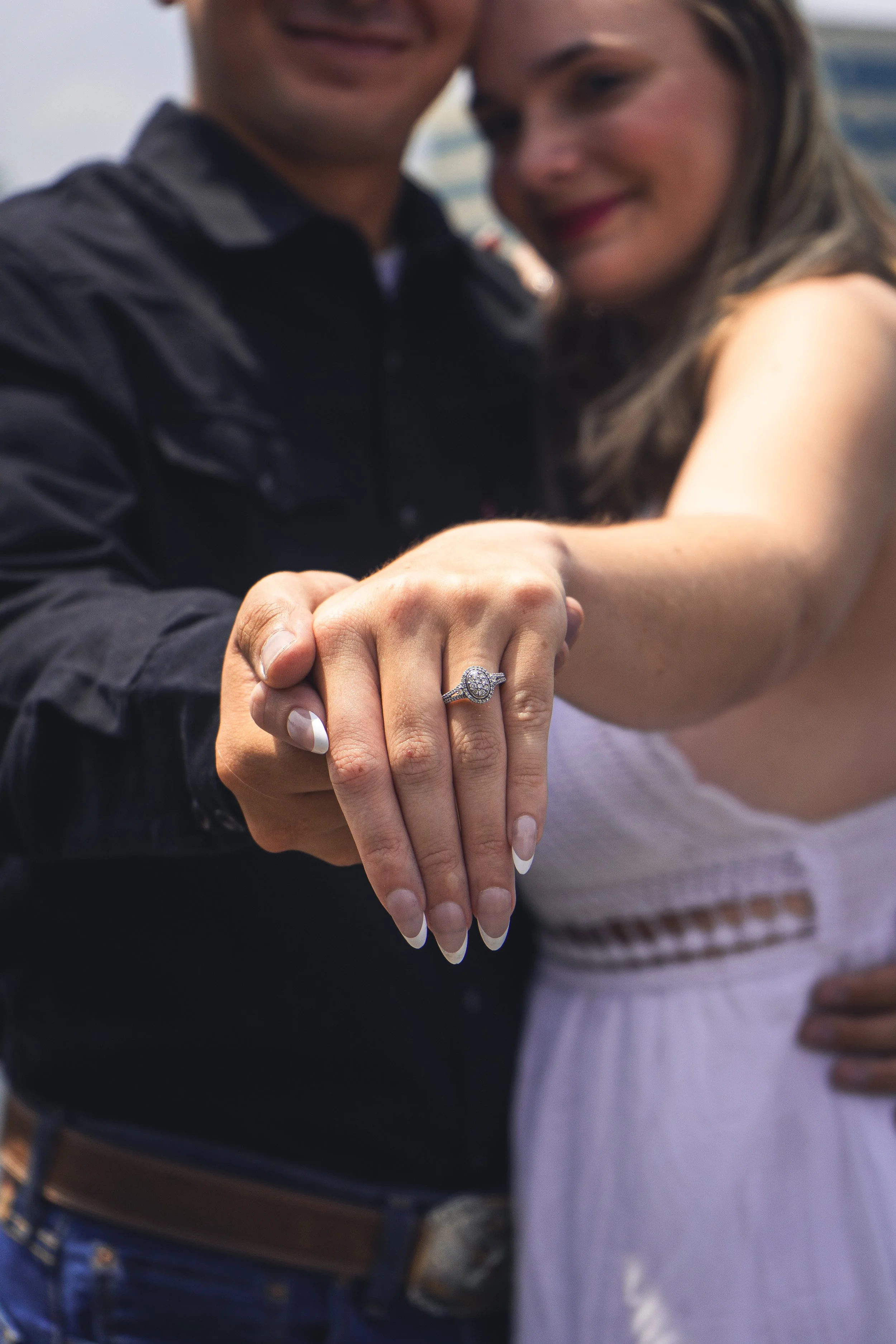Close-up of a couple holding hands, showcasing a diamond engagement ring. The woman has long, manicured nails, and they are smiling. The background suggests an outdoor setting.