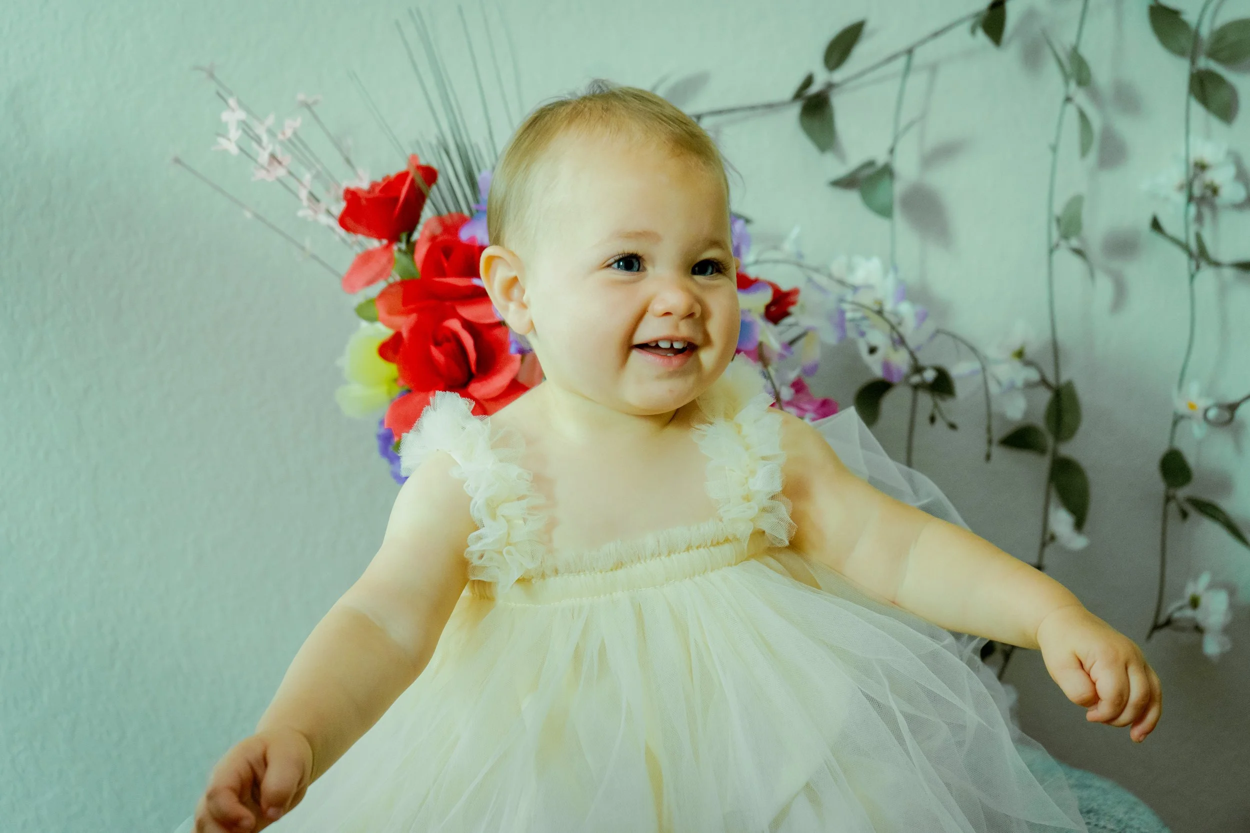 A smiling toddler girl wearing a light yellow dress with ruffled straps, sitting in front of a backdrop decorated with colorful flowers and green leaves.