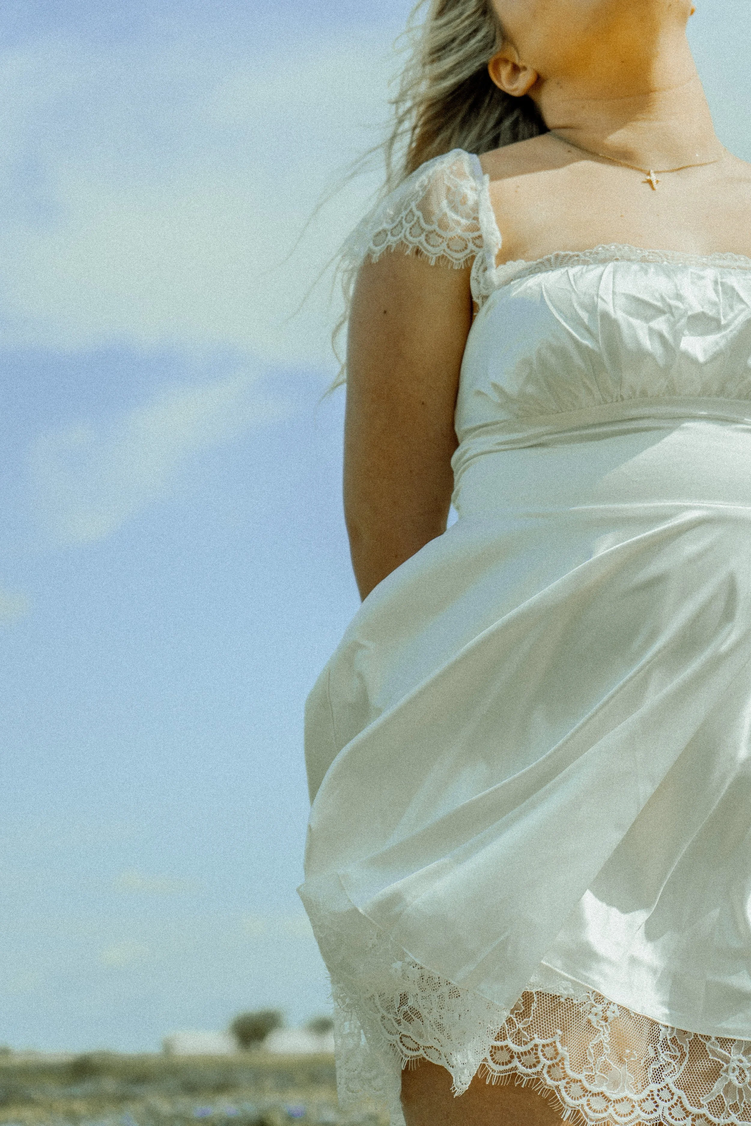 Close-up of a woman wearing a white dress with lace details, standing outdoors with a blurred landscape background.