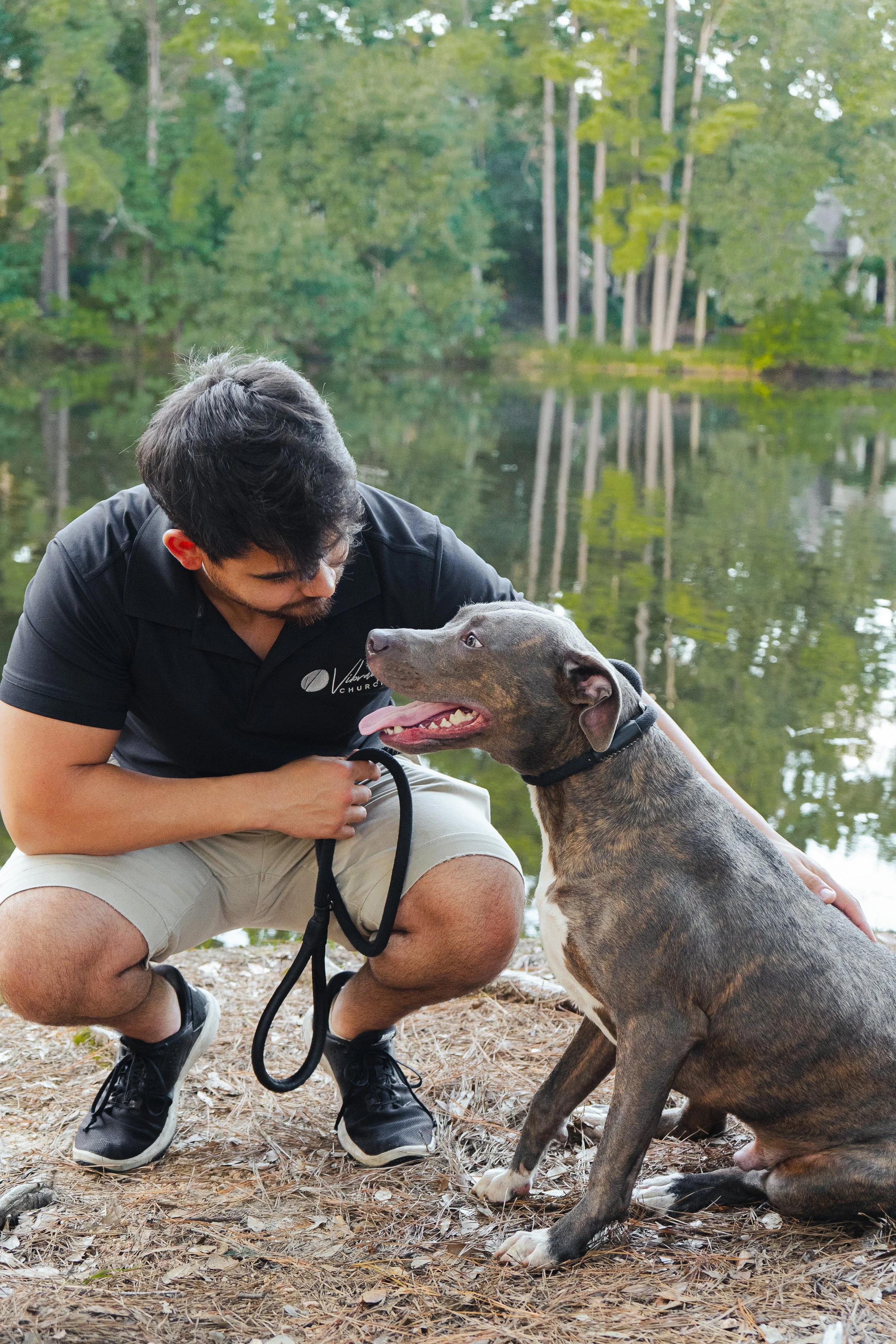 A man with dark hair and a beard crouching on the ground by a lake, holding a black leash attached to a gray and brown dog. The man is wearing a black polo shirt and light-colored shorts. The dog is sitting and looking at the man with its tongue out.