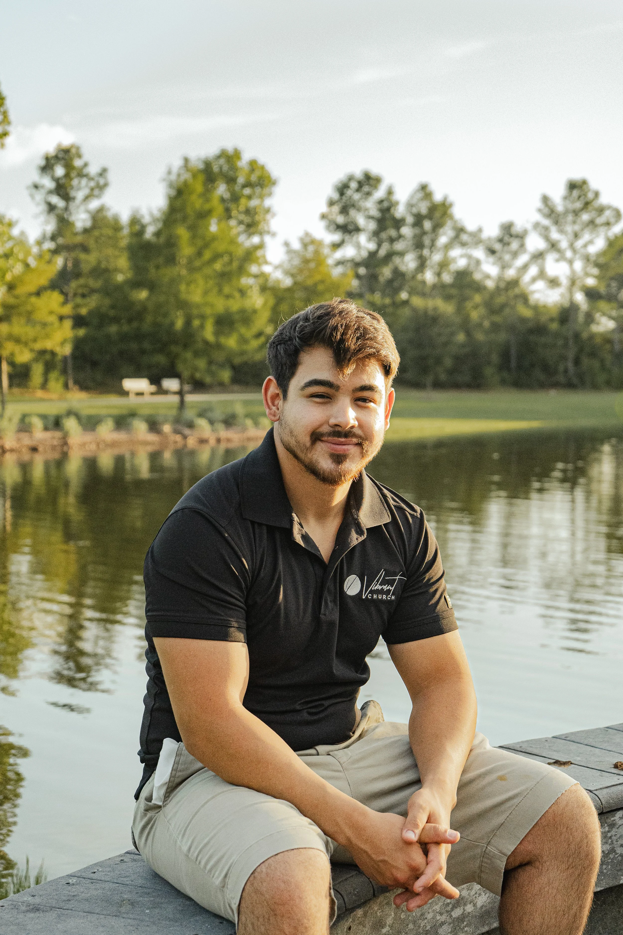 A young man sitting on a dock by a lake in the late afternoon, smiling at the camera, wearing a black polo shirt and beige shorts, with trees and benches in the background.