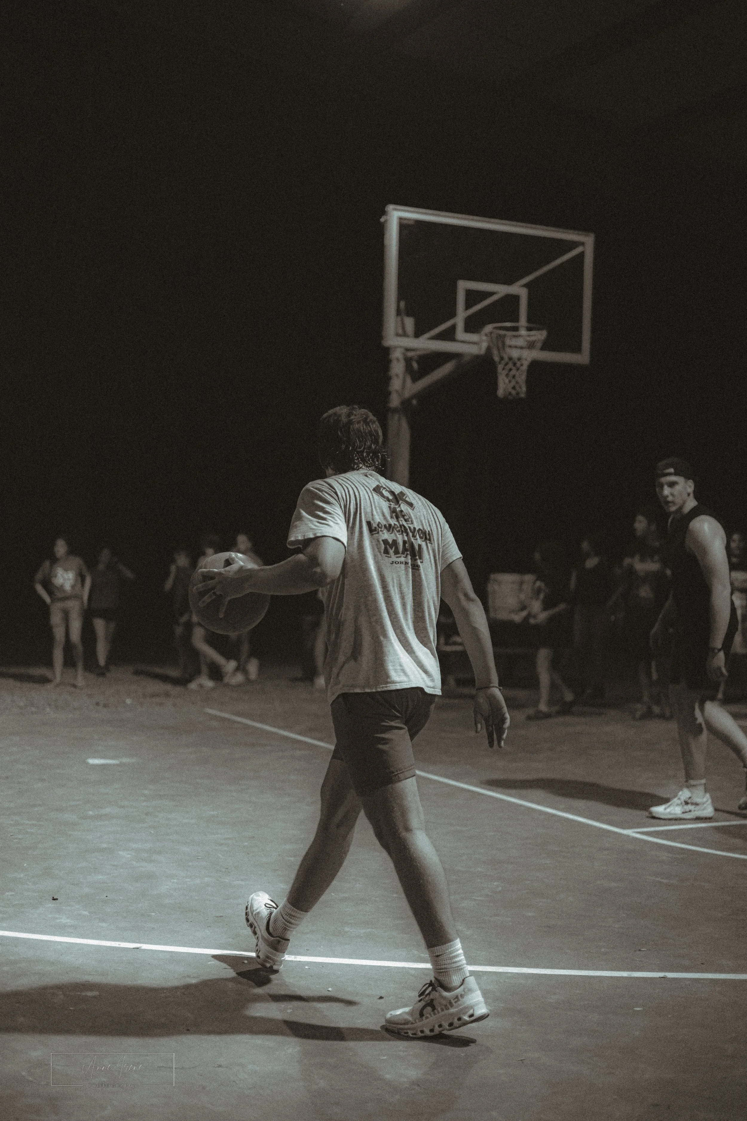 Young man playing basketball on an outdoor court at night, surrounded by other players and spectators.