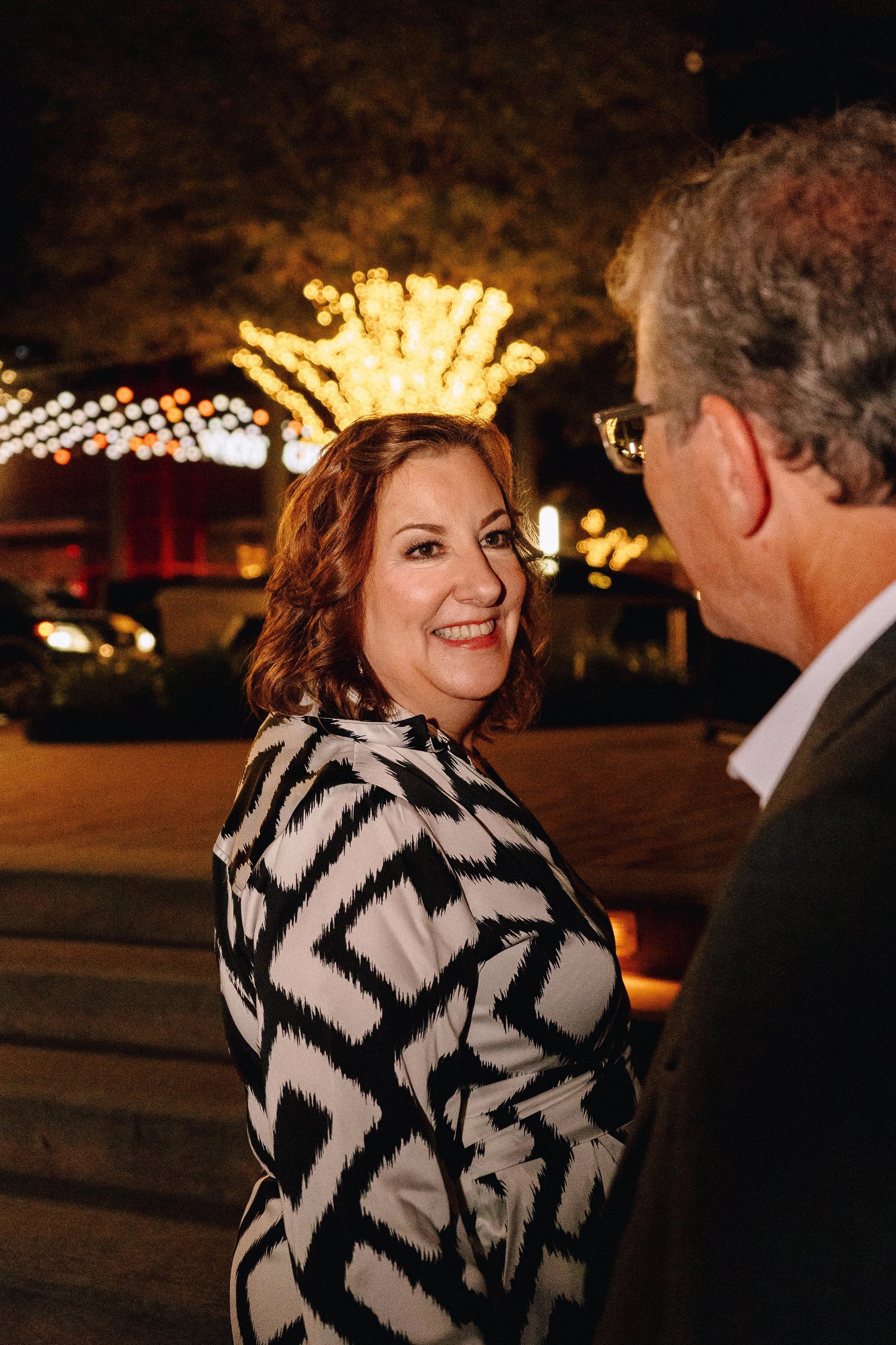A woman with red hair in a black and white patterned dress smiling at a man wearing glasses, in a night-time outdoor setting with festive lights and blurred background.