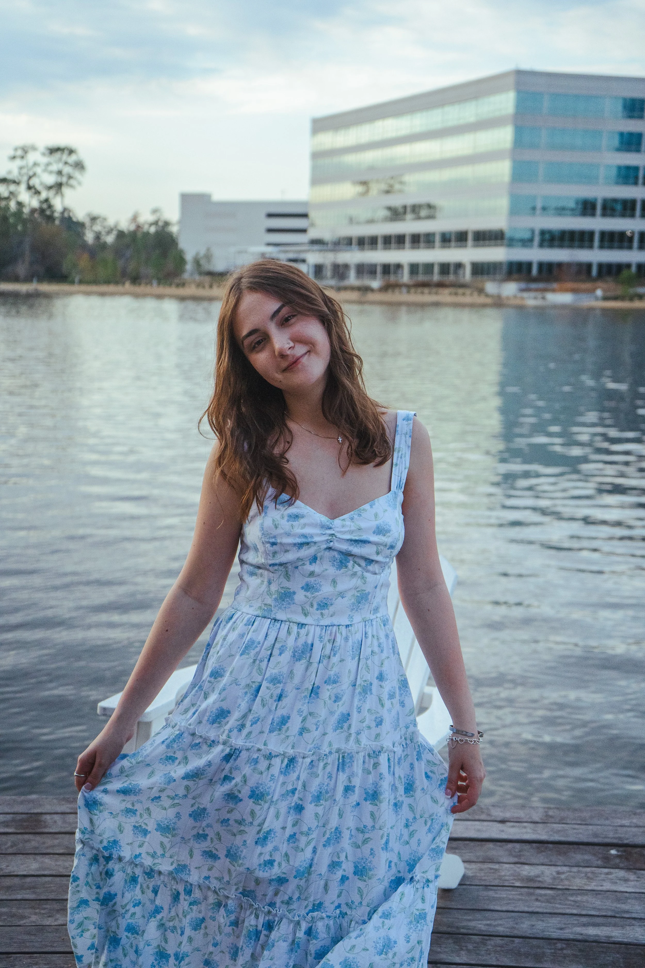 A young woman in a light blue floral dress stands on a wooden dock by a body of water, smiling softly. In the background, there are modern glass buildings and some trees under a cloudy sky.