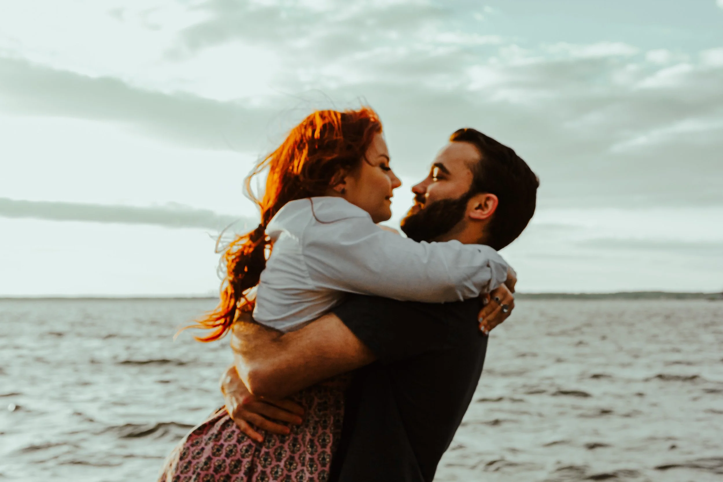 A couple embracing at the beach during sunset, with the woman being lifted by the man, both smiling and looking at each other.