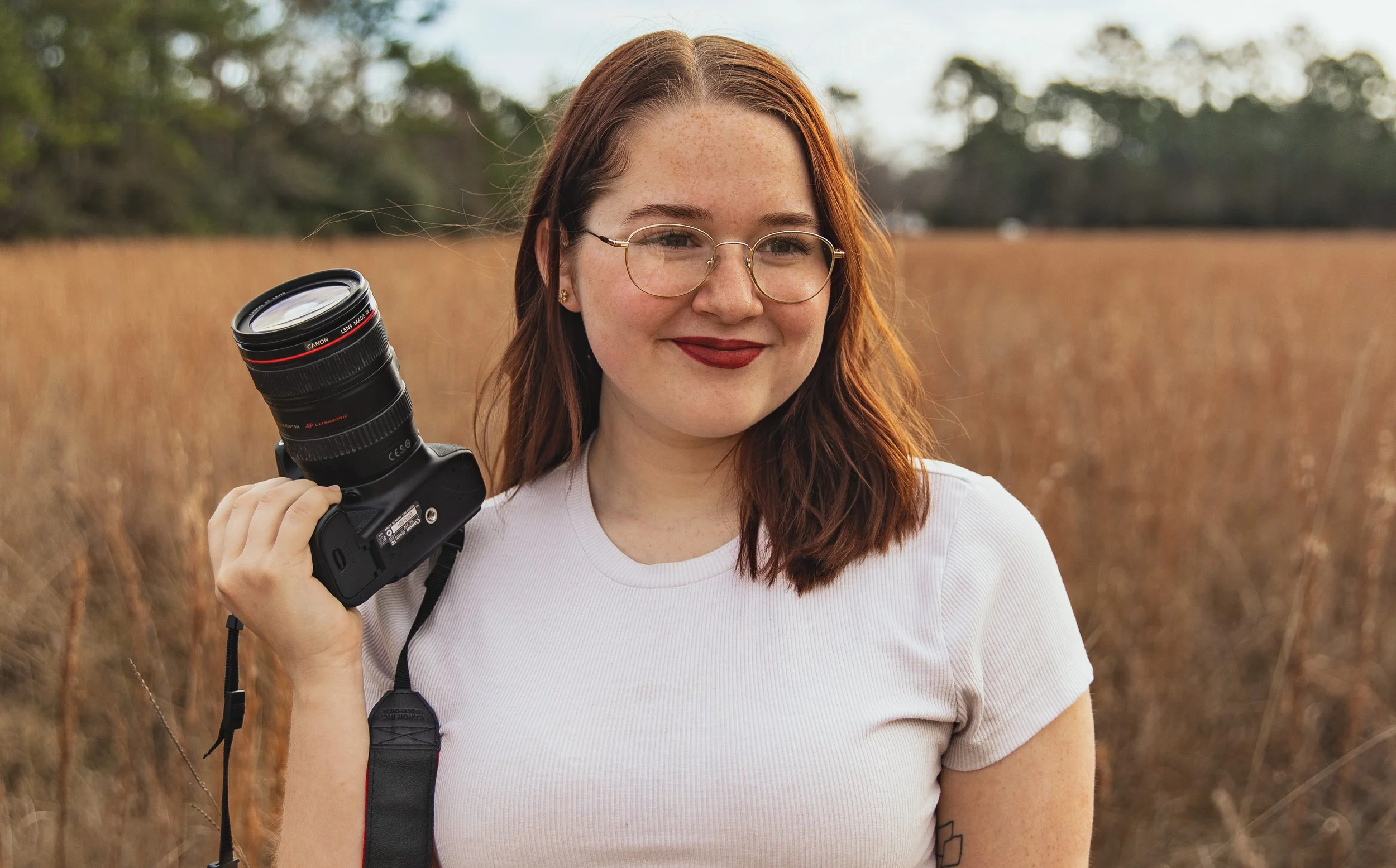 A woman with red hair, glasses, and freckles holds a camera with a large lens on her shoulder outdoors in a field of tall, dry grass at sunset.