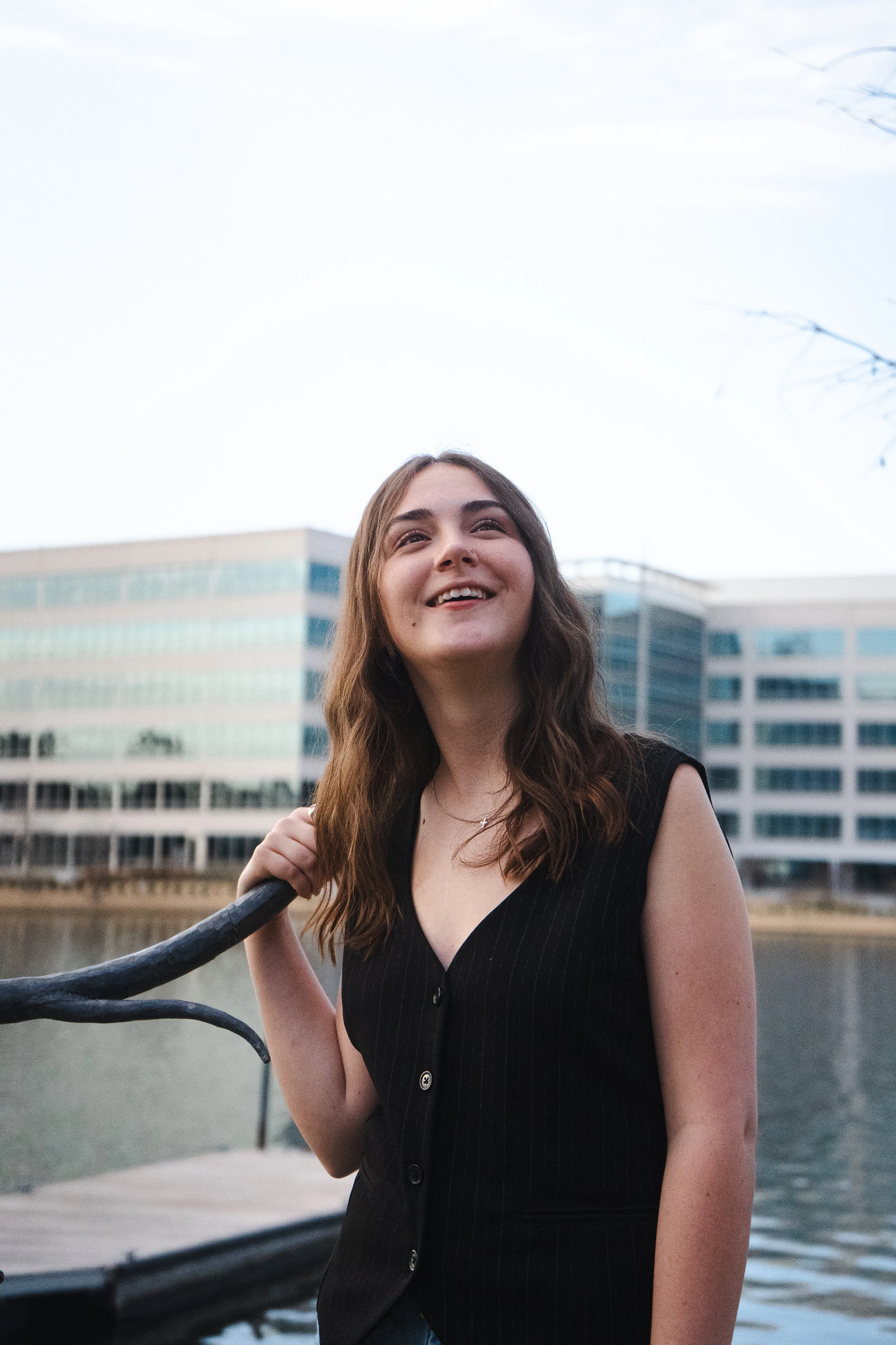 A young woman with long brown hair smiling while standing on a boat by a river with modern buildings in the background.