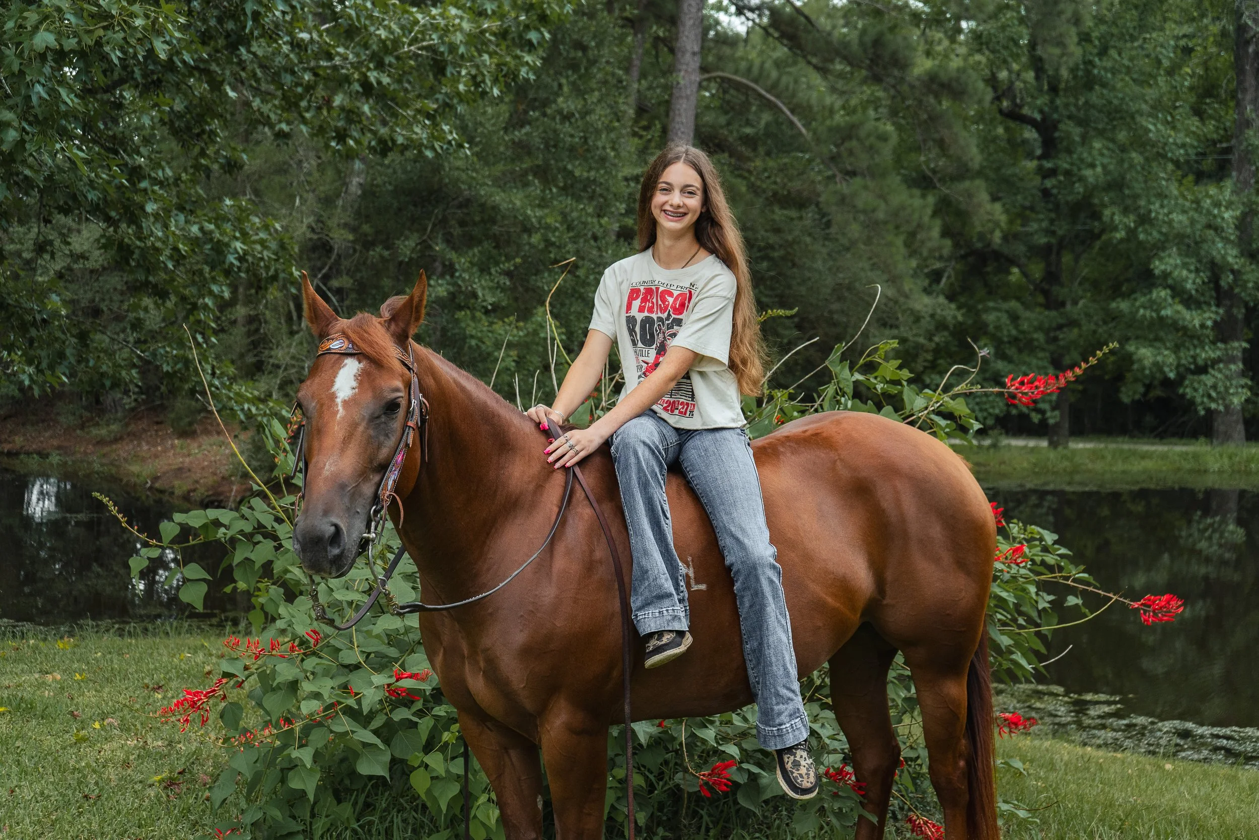 A smiling young girl with long brown hair riding a chestnut horse through a green wooded outdoor area with red flowers and water nearby.