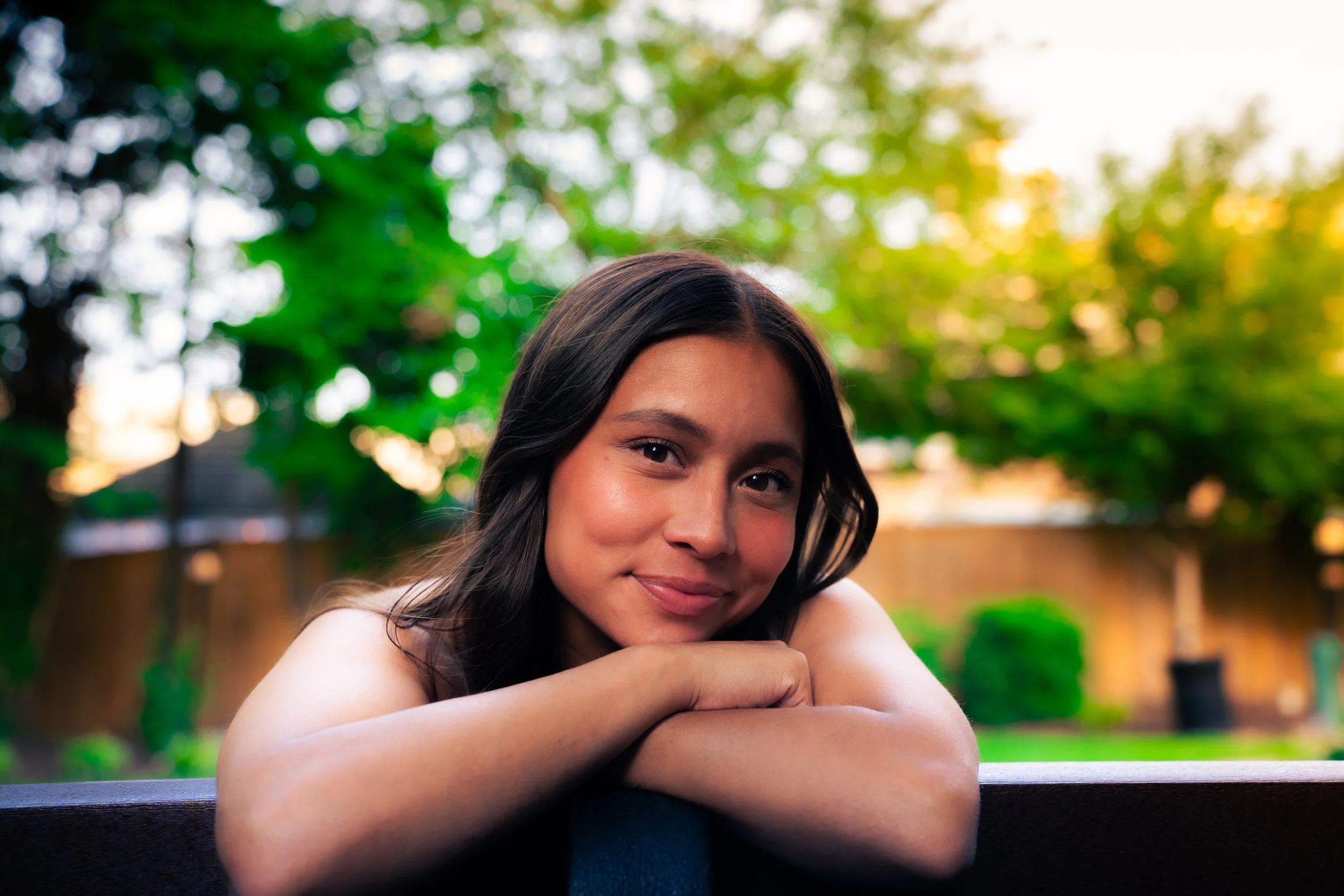 A young woman with dark hair and a light complexion resting her head on her hands on a bench in a park with green trees and sunlight in the background.