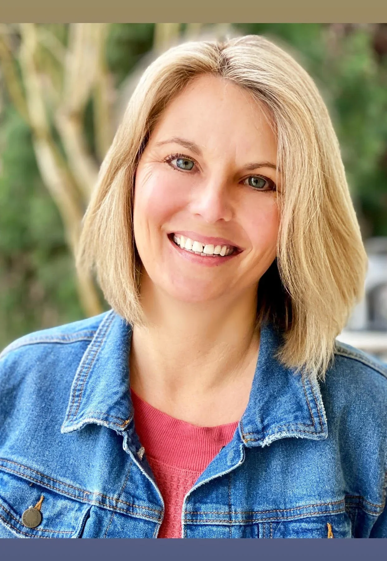 A woman with shoulder-length blonde hair, blue eyes, and a wide smile showing teeth. She is wearing a red top and a blue denim jacket, with a blurred outdoor green and yellow background.