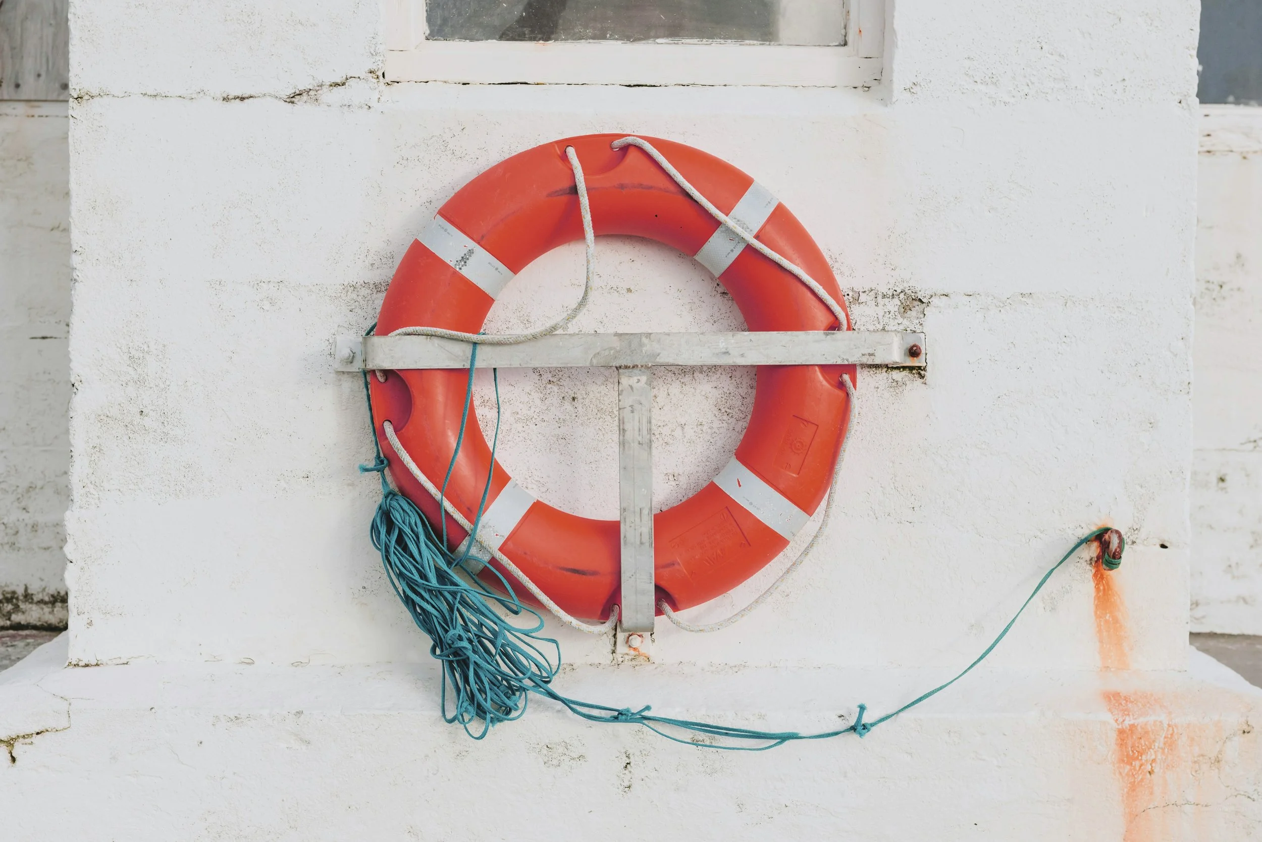 Orange life preserver with white reflectors mounted on a white stucco wall, with blue and green ropes hanging down.