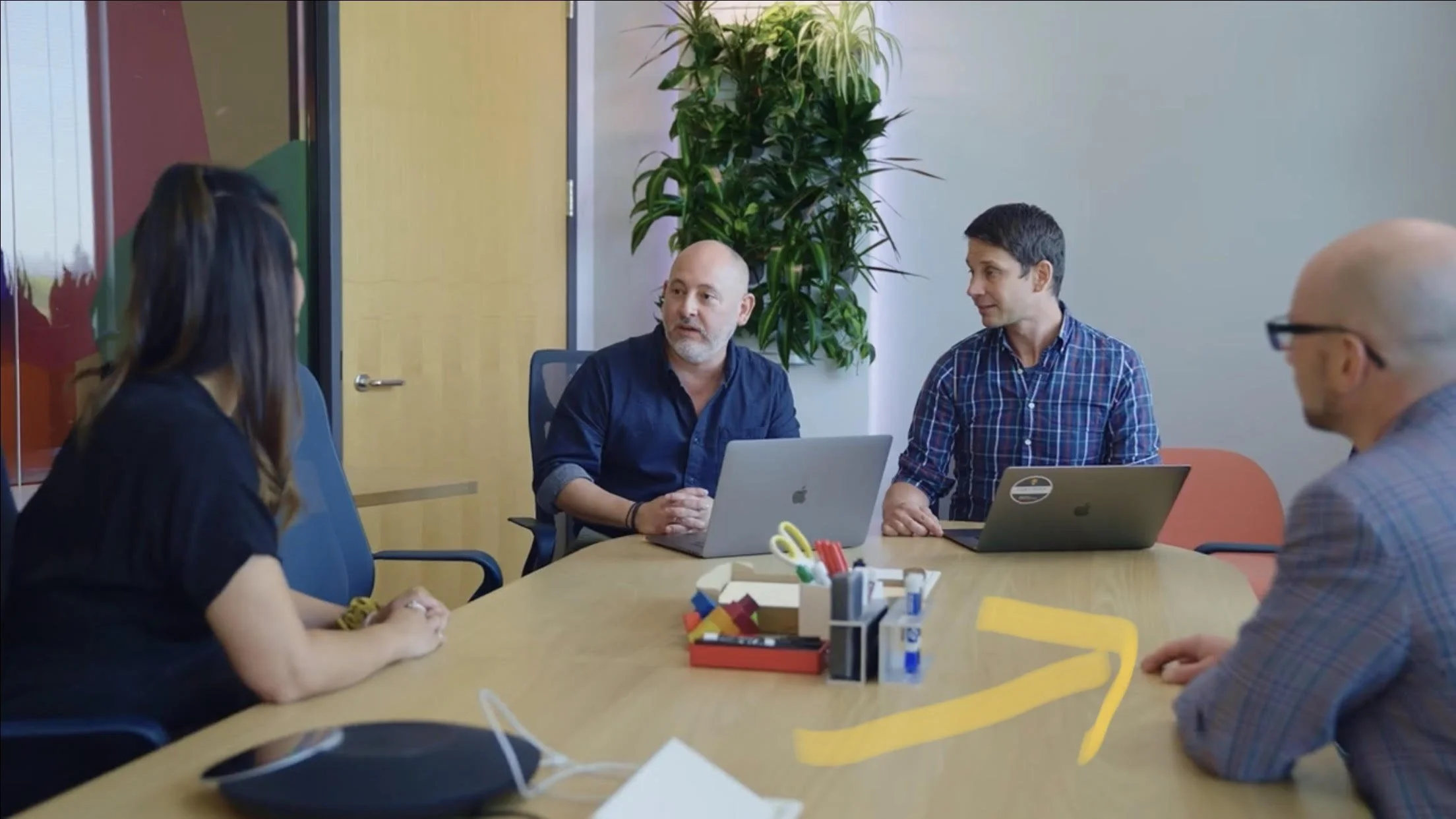 Four people sitting around a conference table in a meeting room with laptops, pens, and office supplies. One woman with long dark hair faces a man with short dark hair wearing a plaid shirt, and another man with a shaved head and glasses faces a woman with long hair. There is a large plant and closed door in the background.