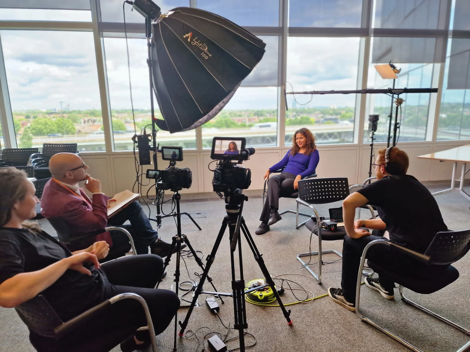 A woman in a blue shirt seated in front of a camera with lighting equipment, participating in a video recording or interview in a room with large windows looking out over a cityscape.