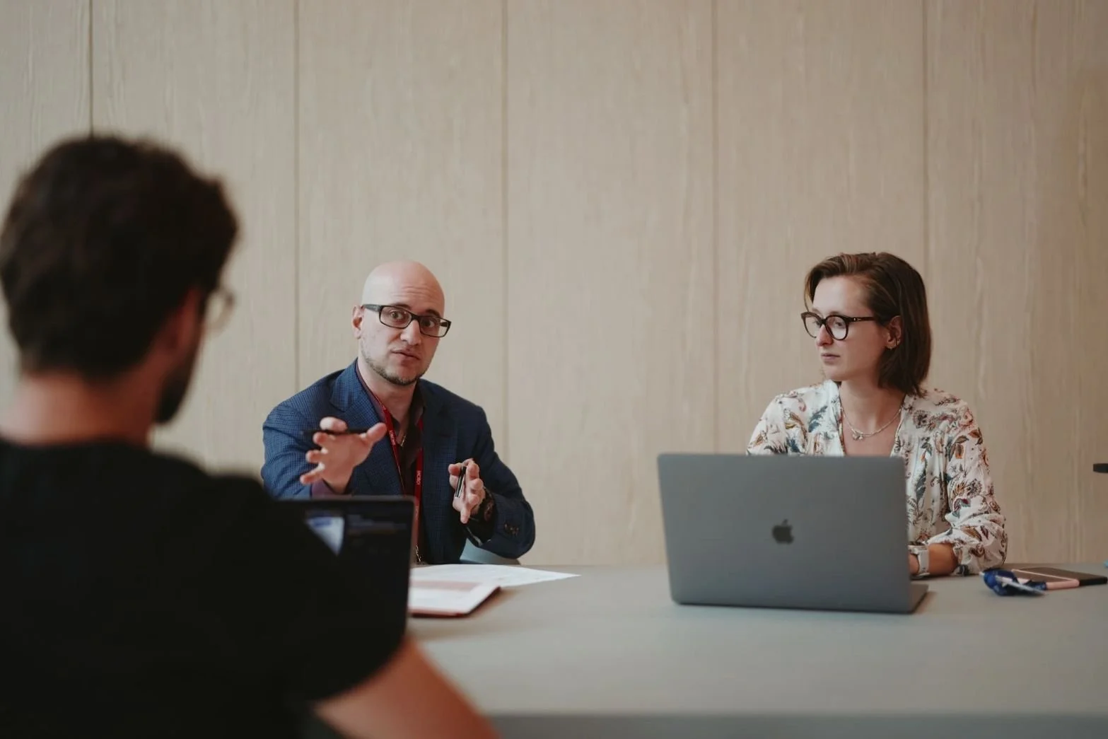 Three people sitting at a conference table during a meeting. Two women and one man are engaged in a discussion, with the man speaking and gesturing with his hands. The woman on the right has a laptop in front of her, and the back of the head of the man on the left is visible.