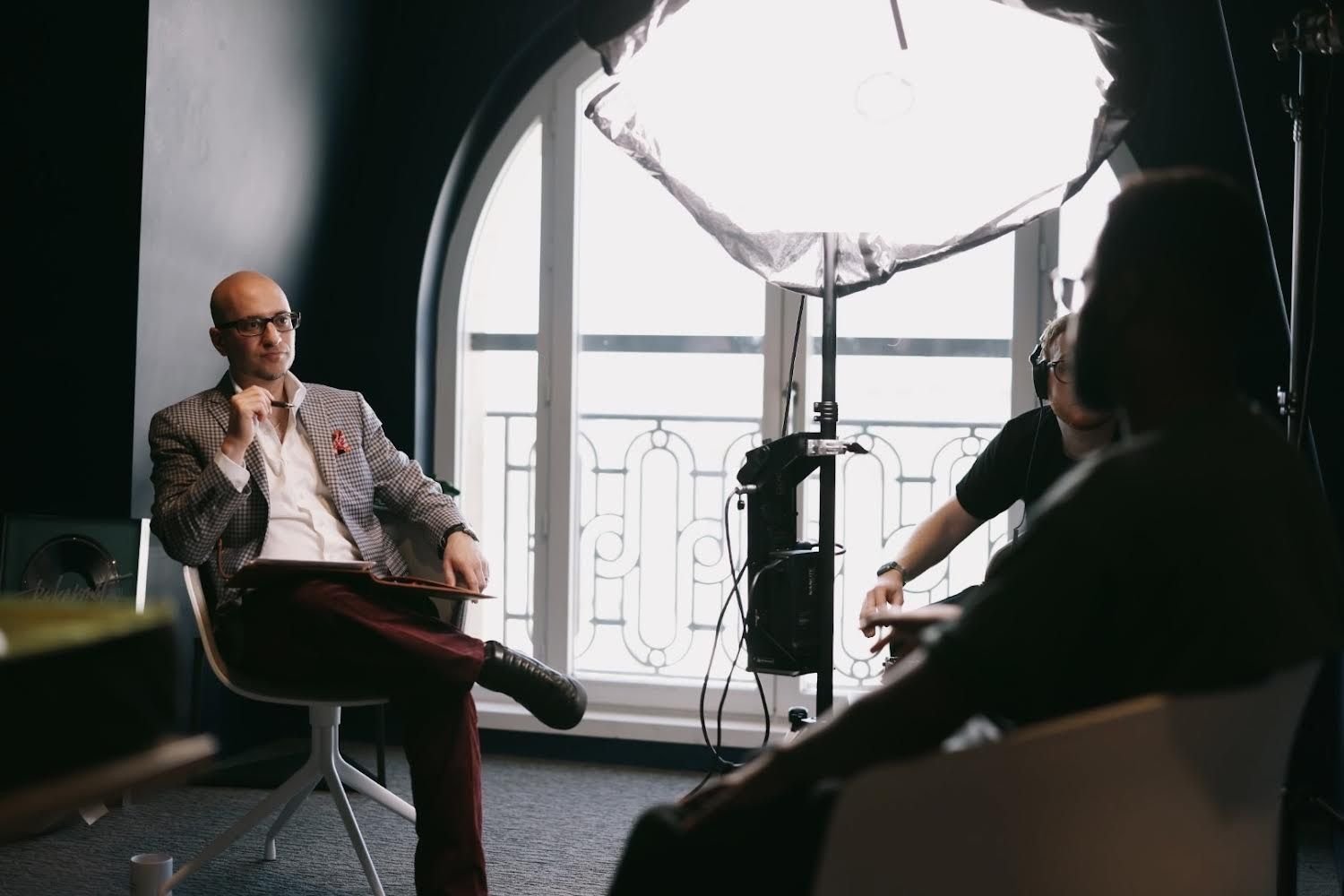 Man in a checkered blazer and glasses being interviewed by two people in a dimly lit room with a large window and balcony railing in the background.